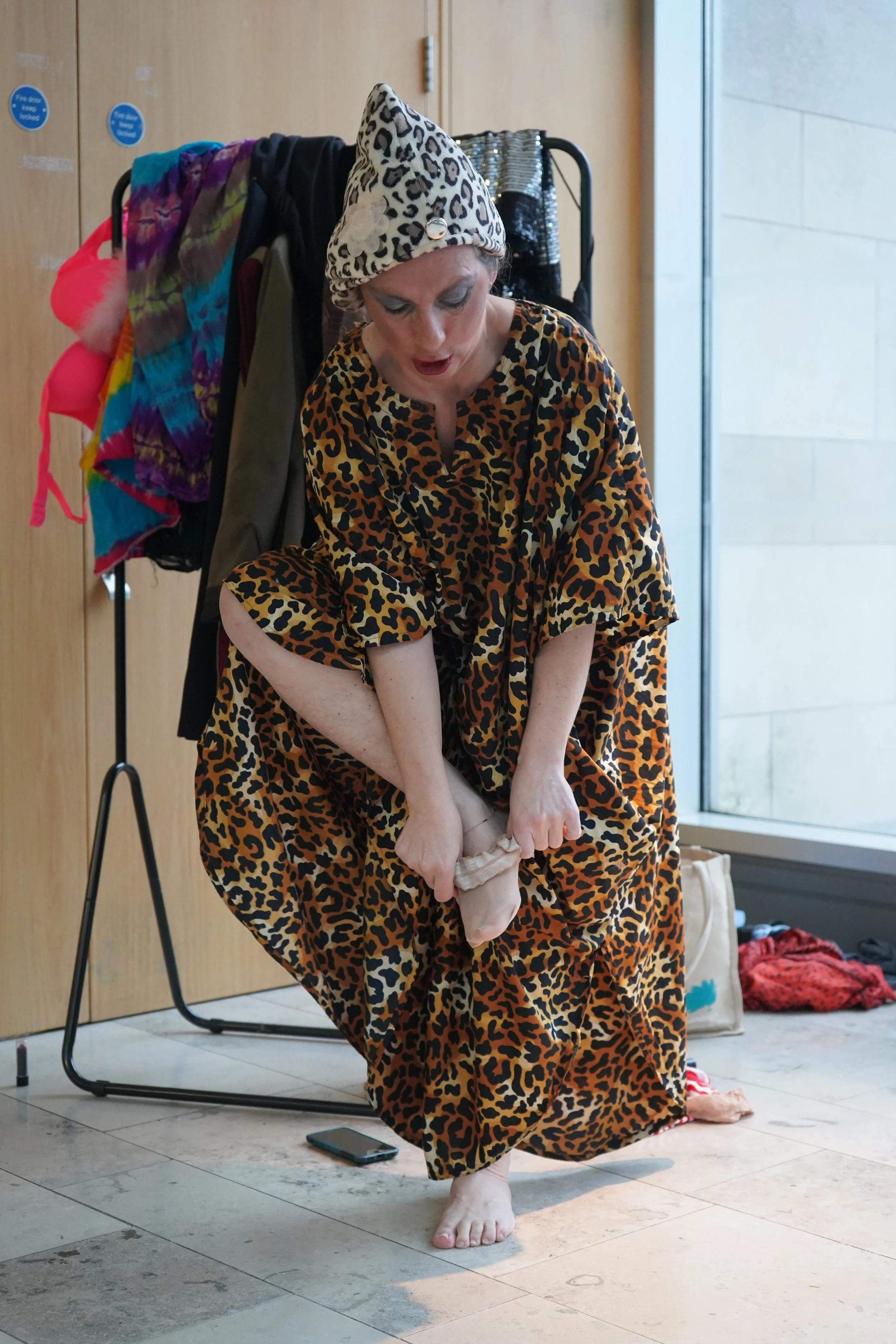 A woman wearing a leopard print dress and hat is performing on the floor, in a performance gesturel act. She has makeup on and is standing on one foot while holding her other leg.