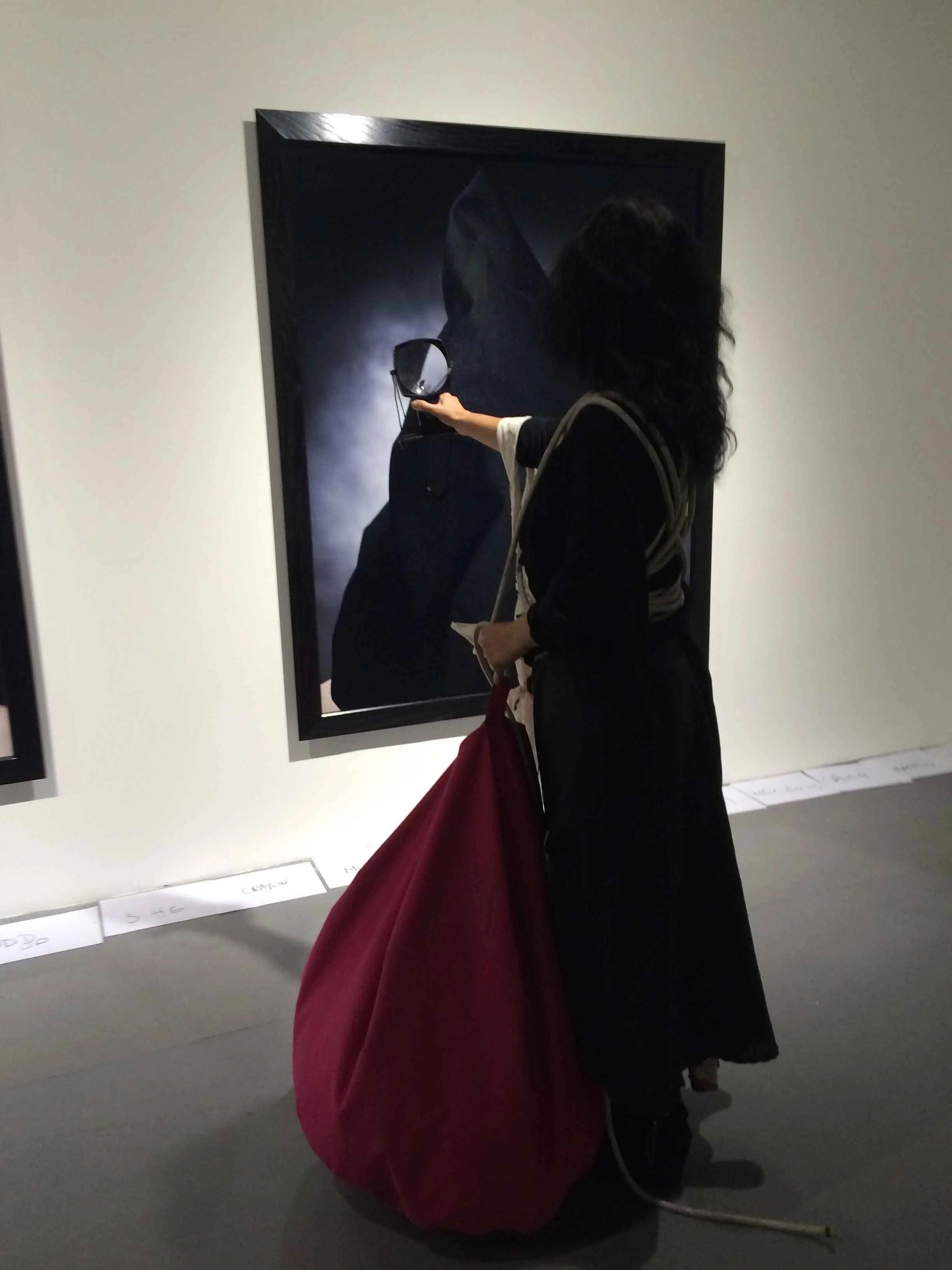 A woman with dark hair, dressed in black, holding a small mirror, standing in front of a large dark artwork and white labels on the wall with handwritten notes at a museum or gallery.