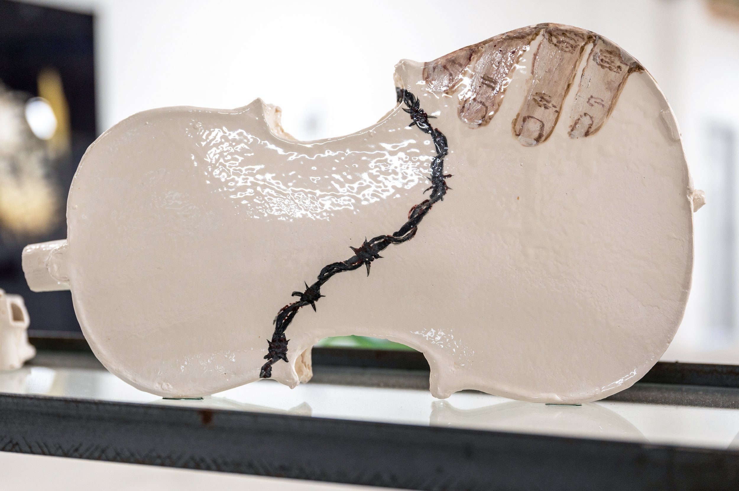 A white ceramic sculpture with a jagged barbed wire drawn running through the middle, with a hand drawn on the edge..