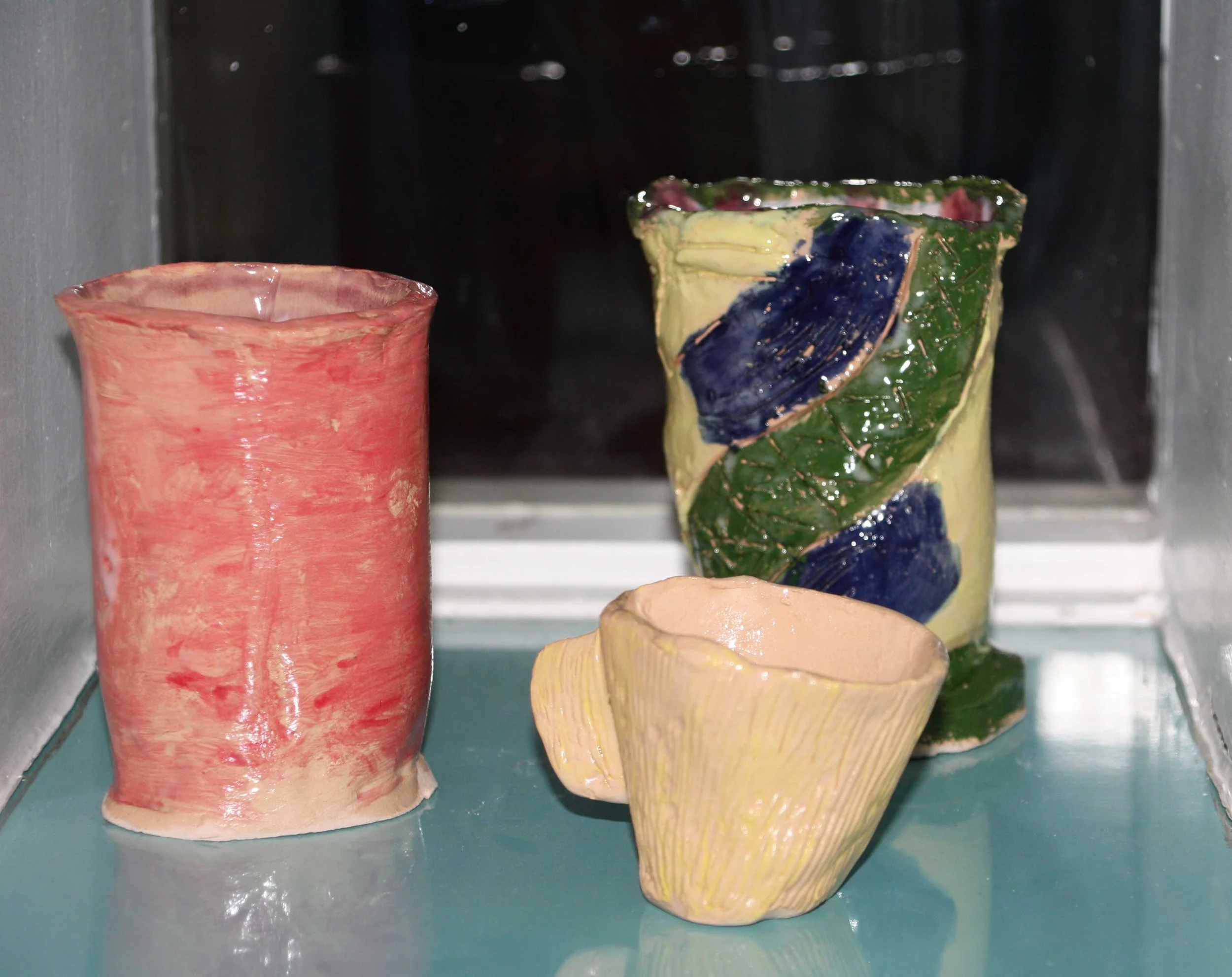 Three ceramic cups with colorful, textured glaze in pink, green, and yellow with blue accents, displayed on a glass shelf.