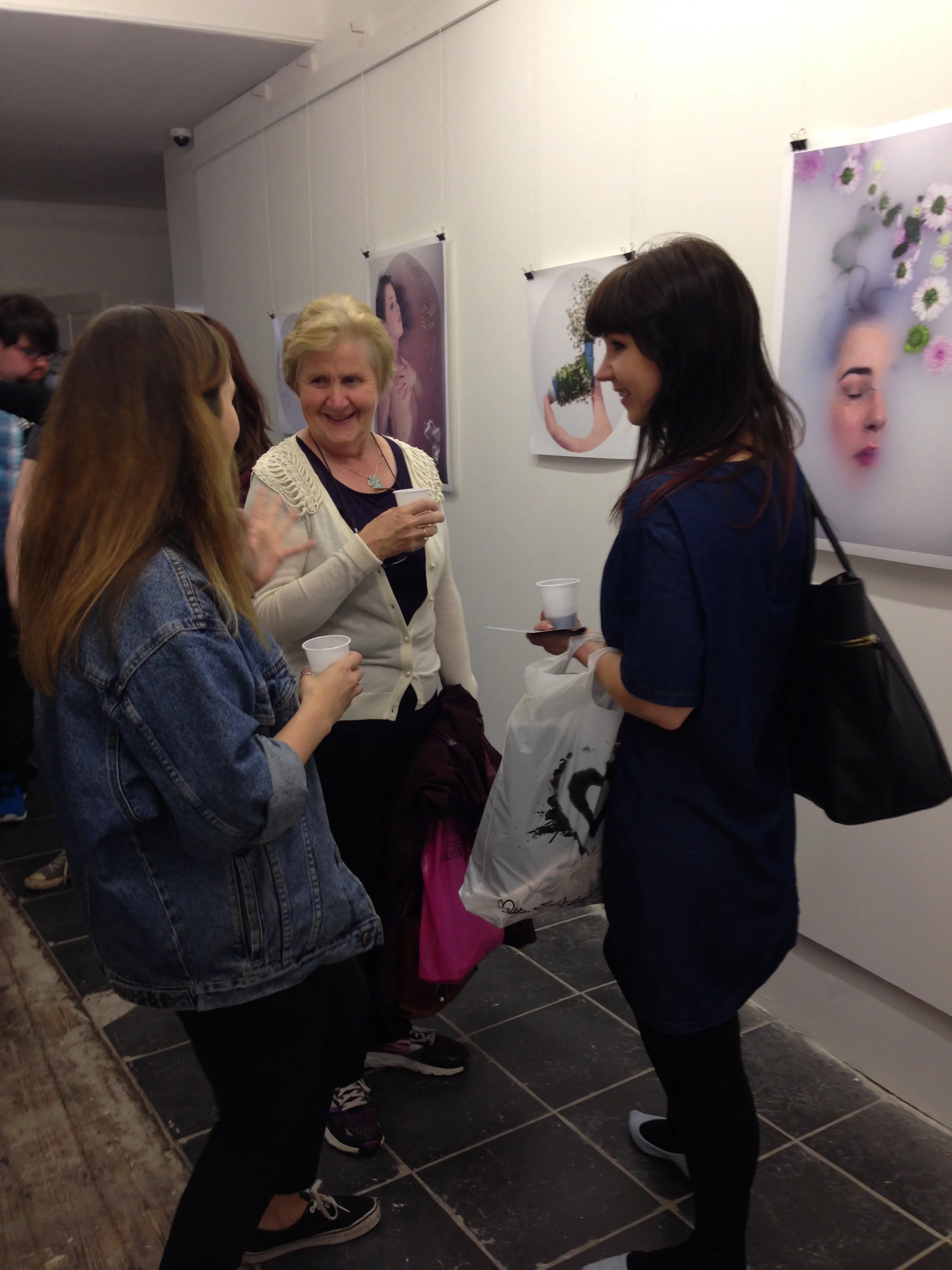 A group of women are conversing at an art gallery exhibition, with photos of women and artistic images hanging on the wall behind them.