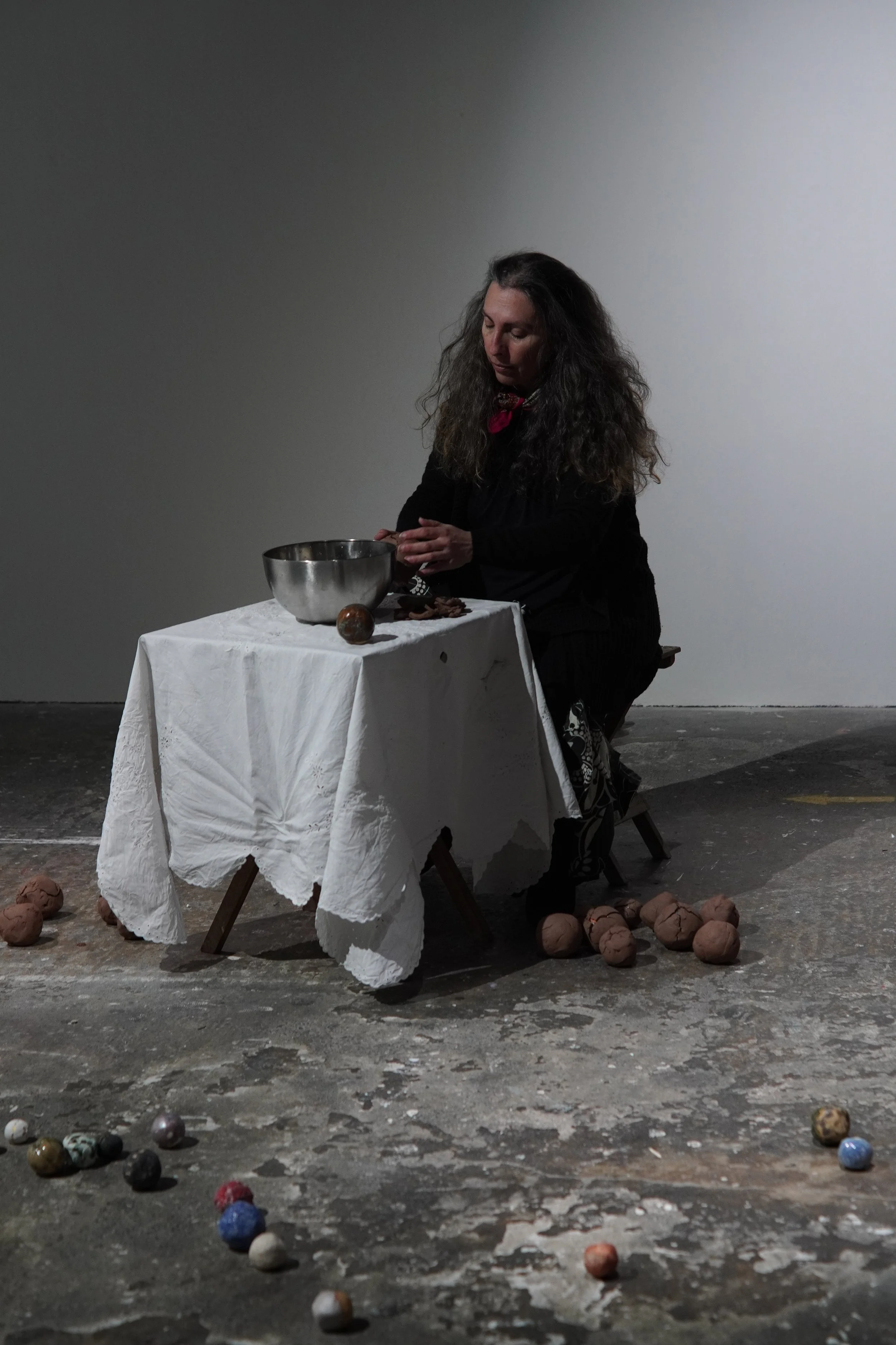 A woman with long, curly hair dressed in black is seated at a small table covered with a white cloth, in a dimly lit room. She appears to be engaged in an activity involving a large metal mixing bowl on the table. The floor is scattered with colorful