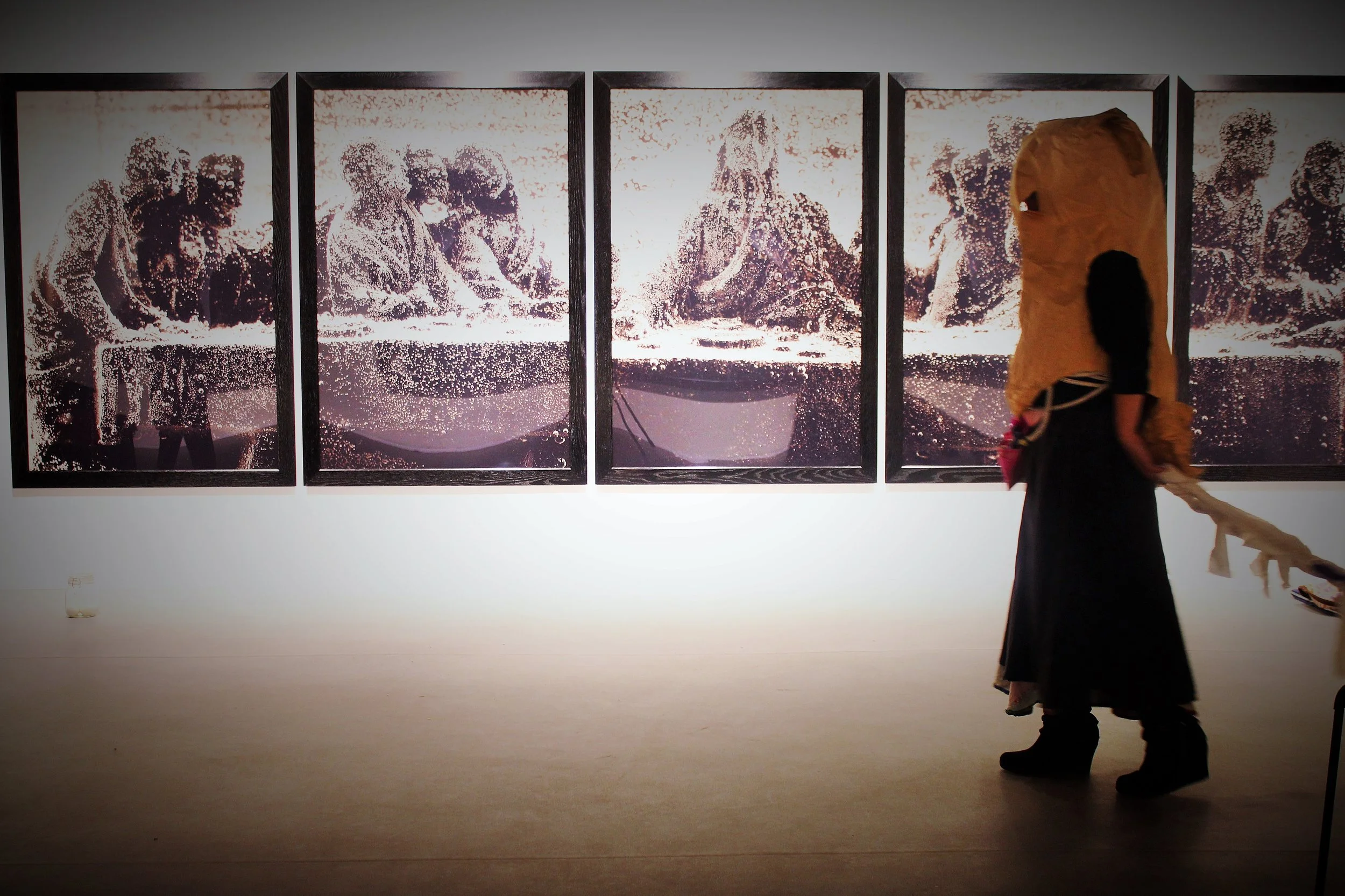 Visitors walking in front of an art installation of three large panels with black and white images of people, with water droplets on the surfaces of the panels.