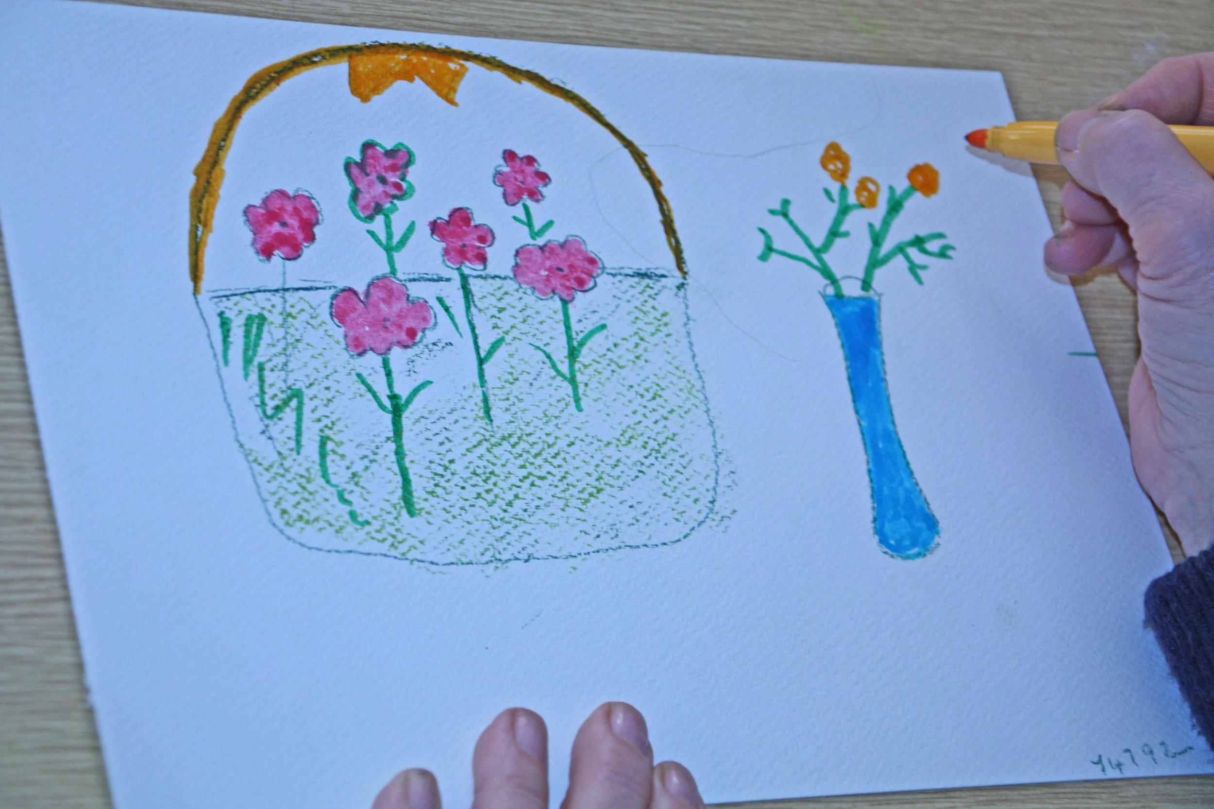 A hand holding a yellow marker is drawing on a piece of paper. The drawing features a flowerpot with pink flowers inside, outlined in green with some inside details. To the right is a blue vase with small orange flowers. The drawing appears to be created with colored markers and crayons.