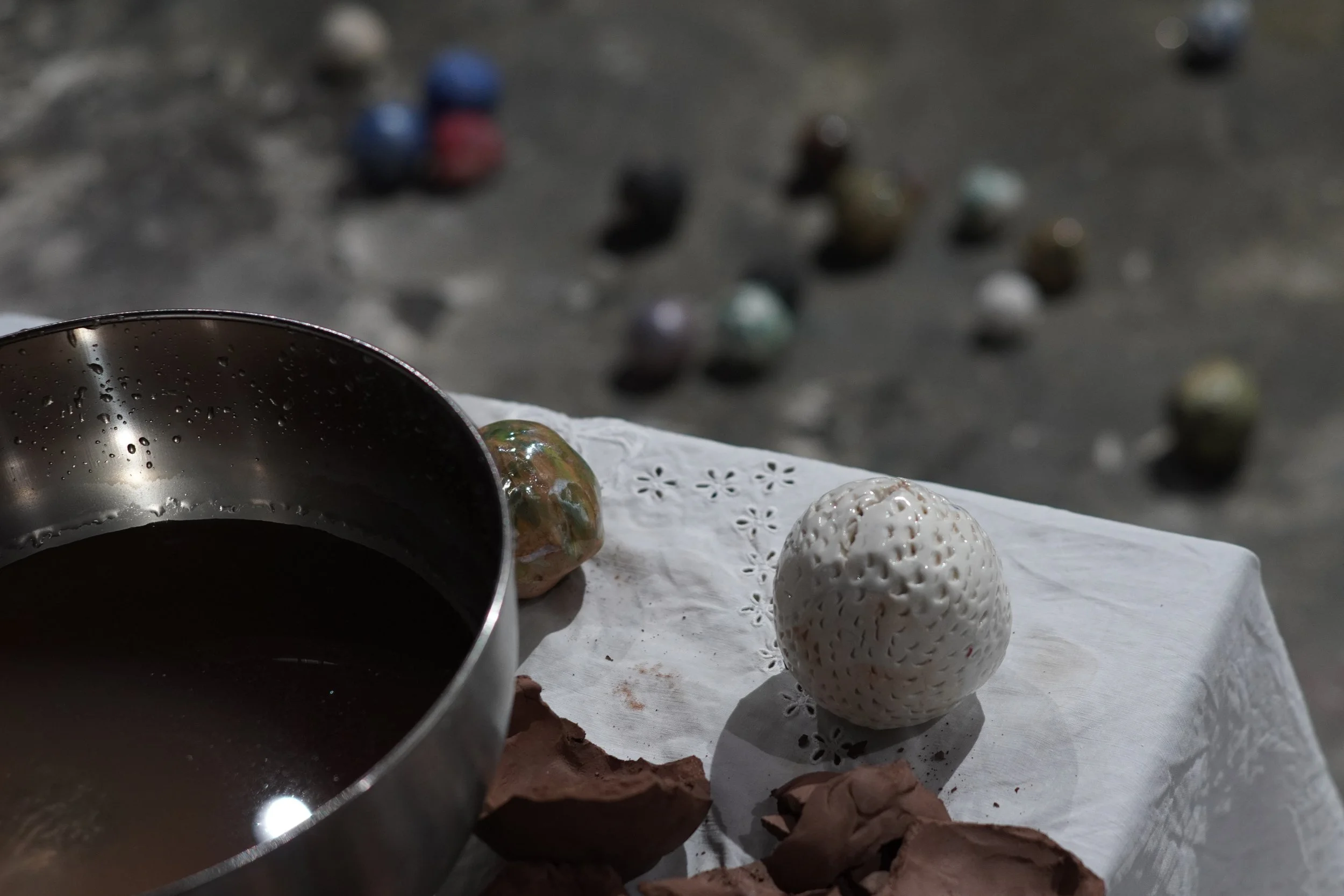 A table covered with a white lace tablecloth holding a stainless steel bowl with water droplets, two decorative white strawberries, and some pieces of broken chocolate. In the background, there are scattered colorful marbles on a dark surface.