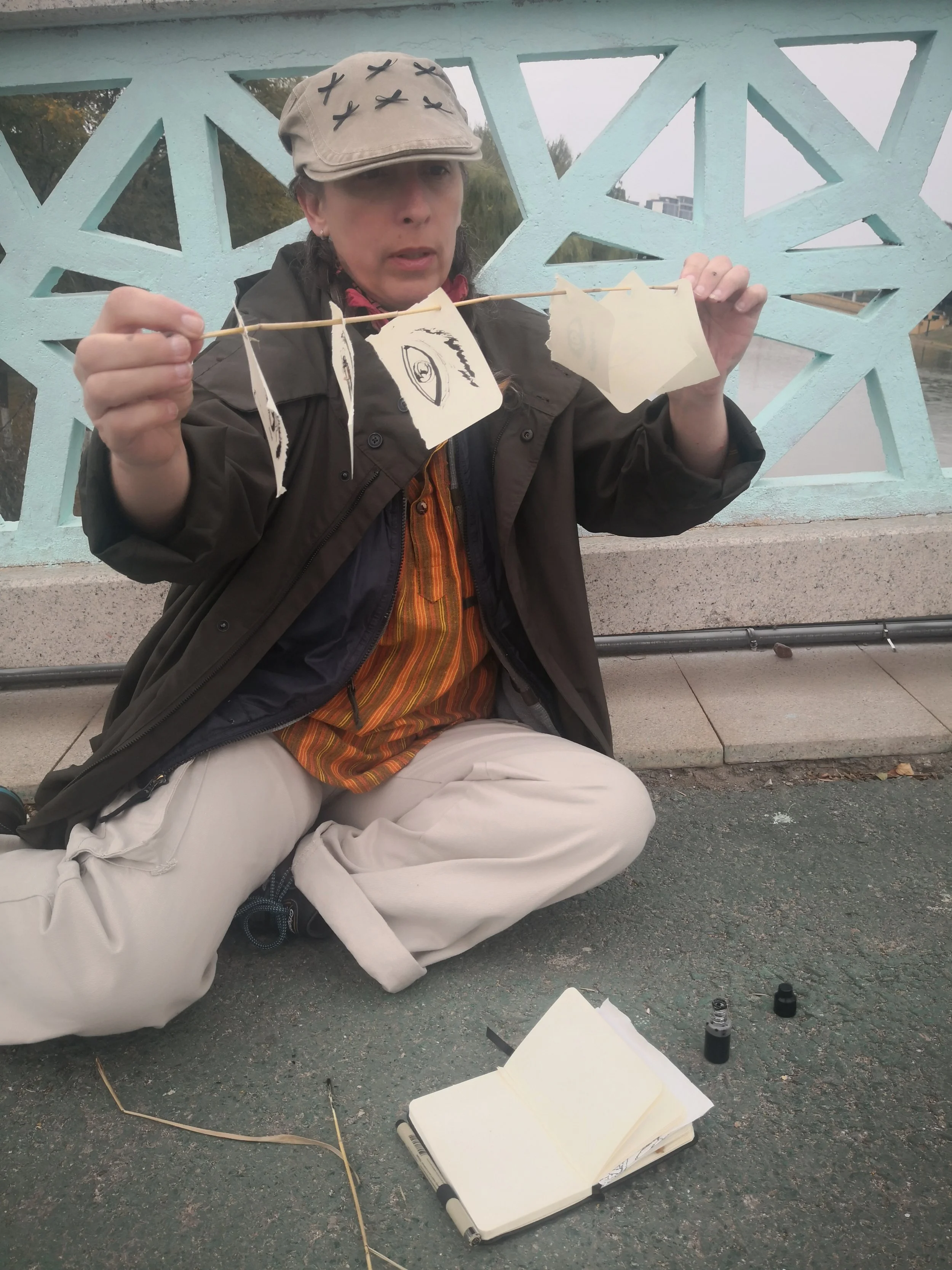 A woman sitting on the ground in front of a decorative teal fence, holding a stick with paper lanterns or decorations. There is an open notebook, marker, and bottles of ink or paint on the ground. She is wearing a beige cap with black X’s, a dark jacket, and beige pants.