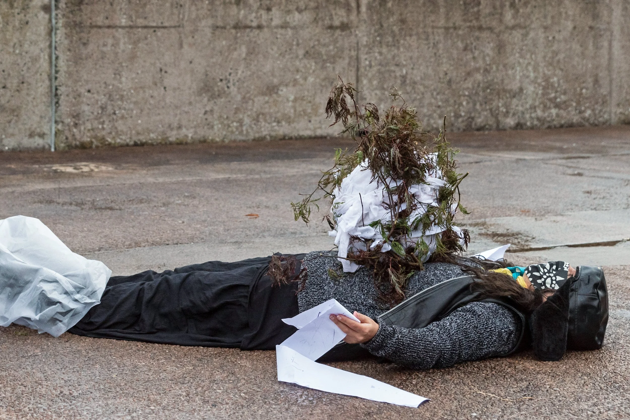 A person lying on the sidewalk with a pile of dead branches and leaves on their face. The person is dressed in dark clothing and is holding papers in their hand.