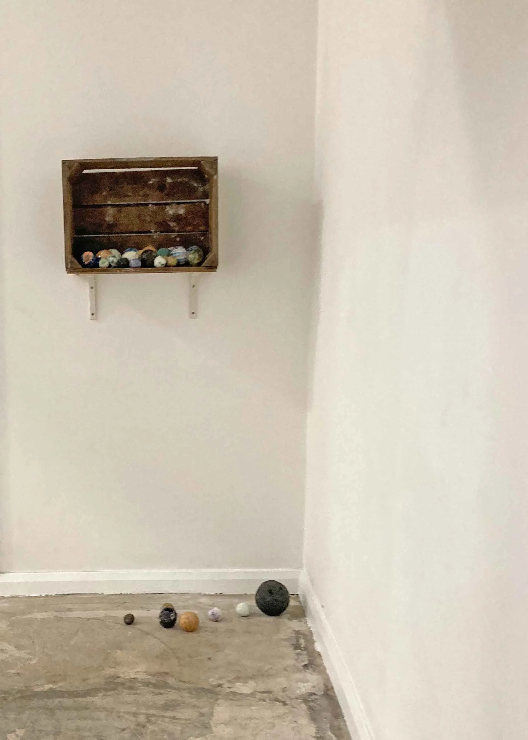 Empty room with white walls, a small wall-mounted wooden shelf with balls of different colors and sizes, and a few balls scattered on the floor.