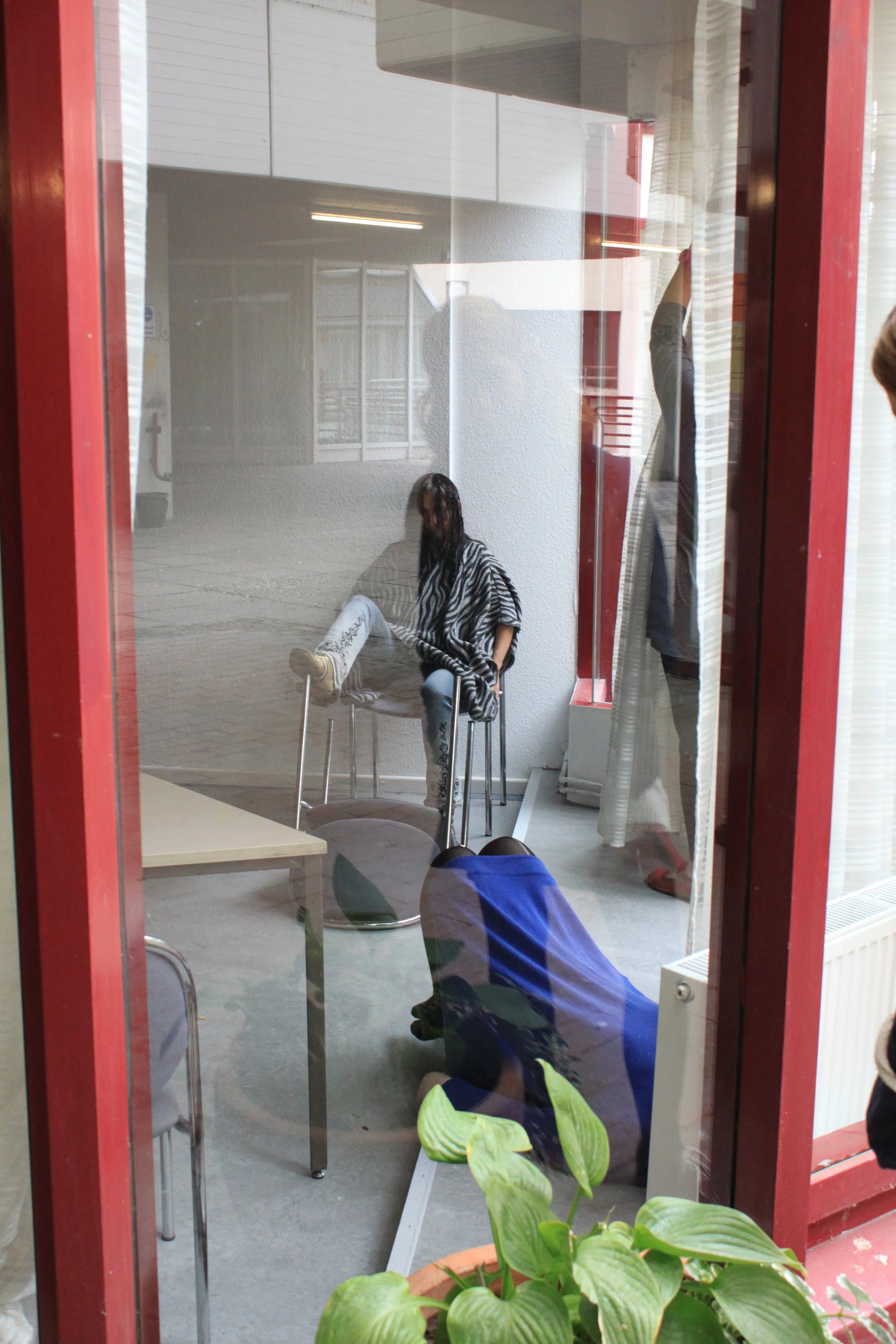 A person in a black and white striped shirt sitting on a chair with water on their face, with other people and chairs visible through a glass window.