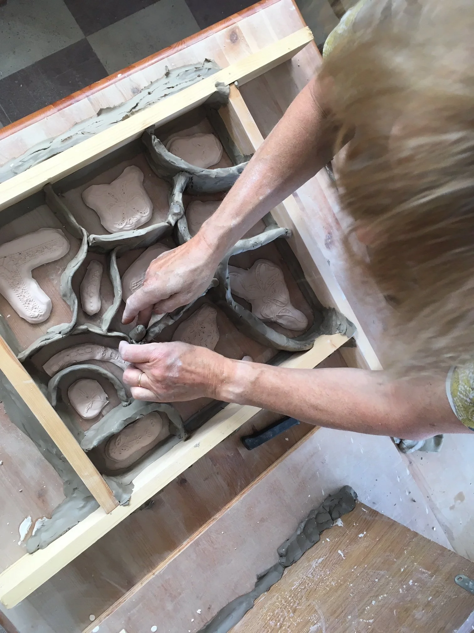 A person is working with clay, using their hands to shape it within a wooden frame. The scene appears to be in an art studio or workshop.