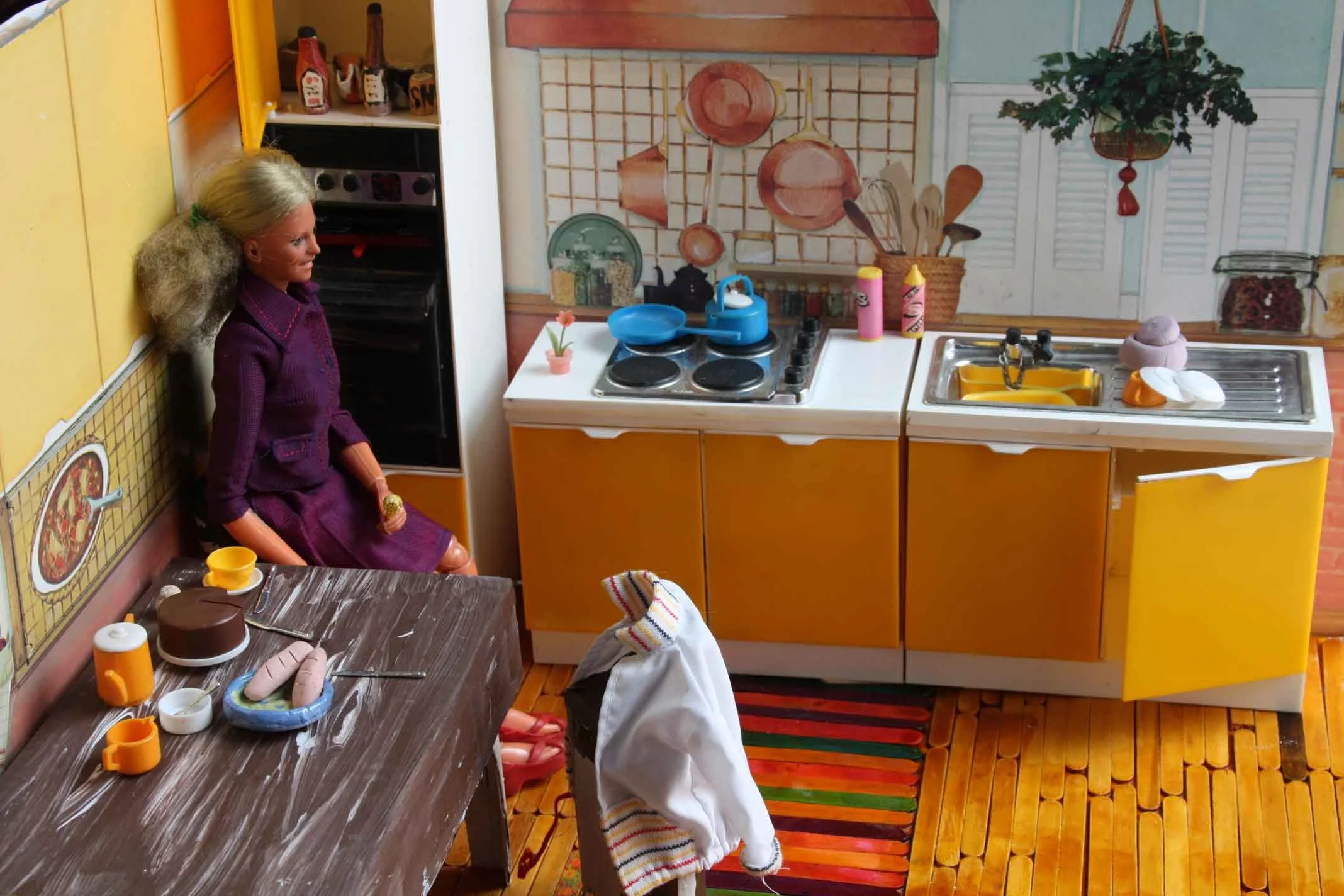 A doll with long blonde hair sitting in a miniature kitchen. The doll is wearing a purple jumpsuit. The kitchen includes a stove with a blue kettle and pots, a sink with dishes, and various kitchen utensils and decorations on the wall.