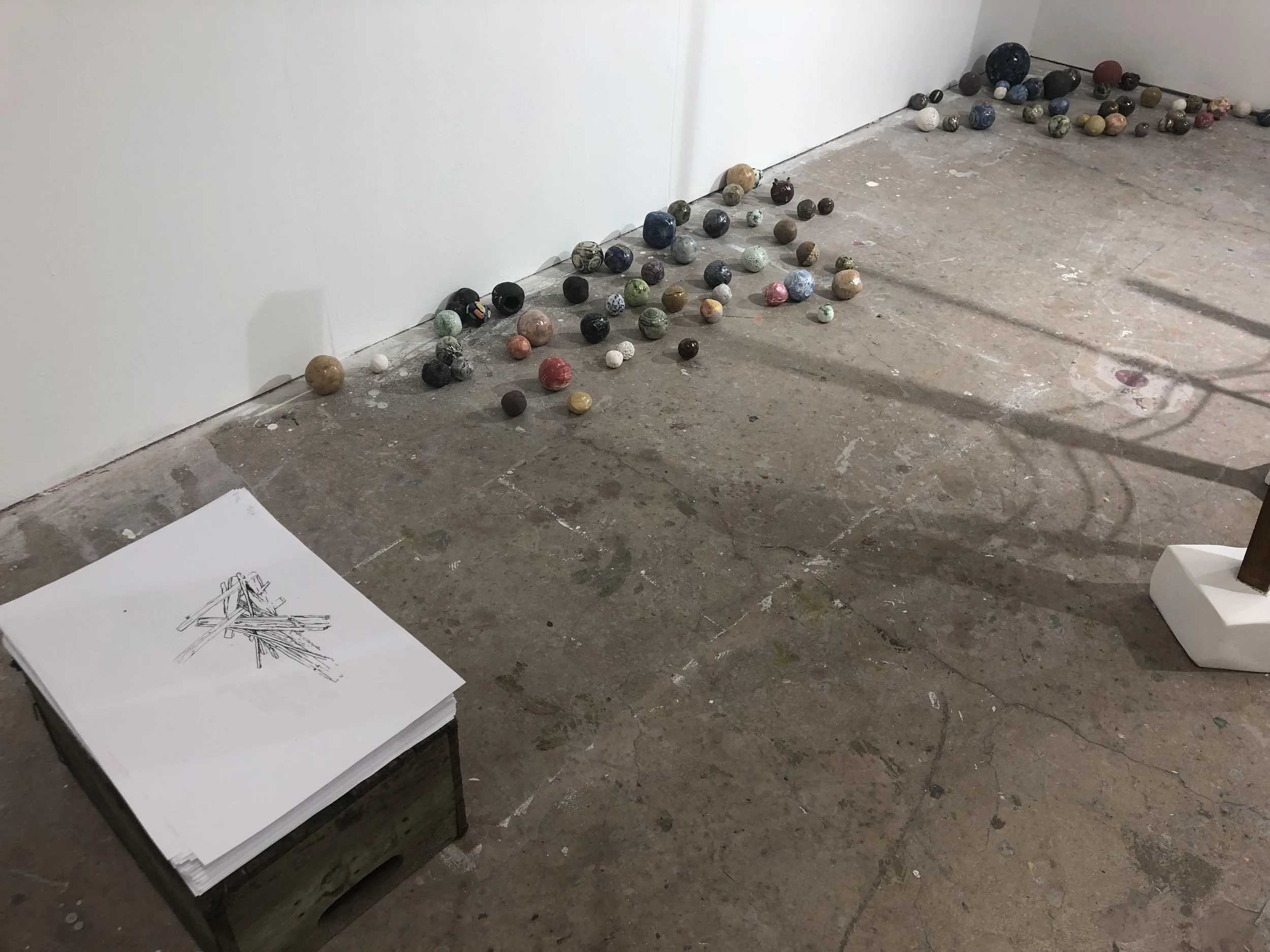 Collection of balls arranged on the floor next to a white wall, with some scattered on the ground, and a sketch pad with architectural drawings on a small table nearby.
