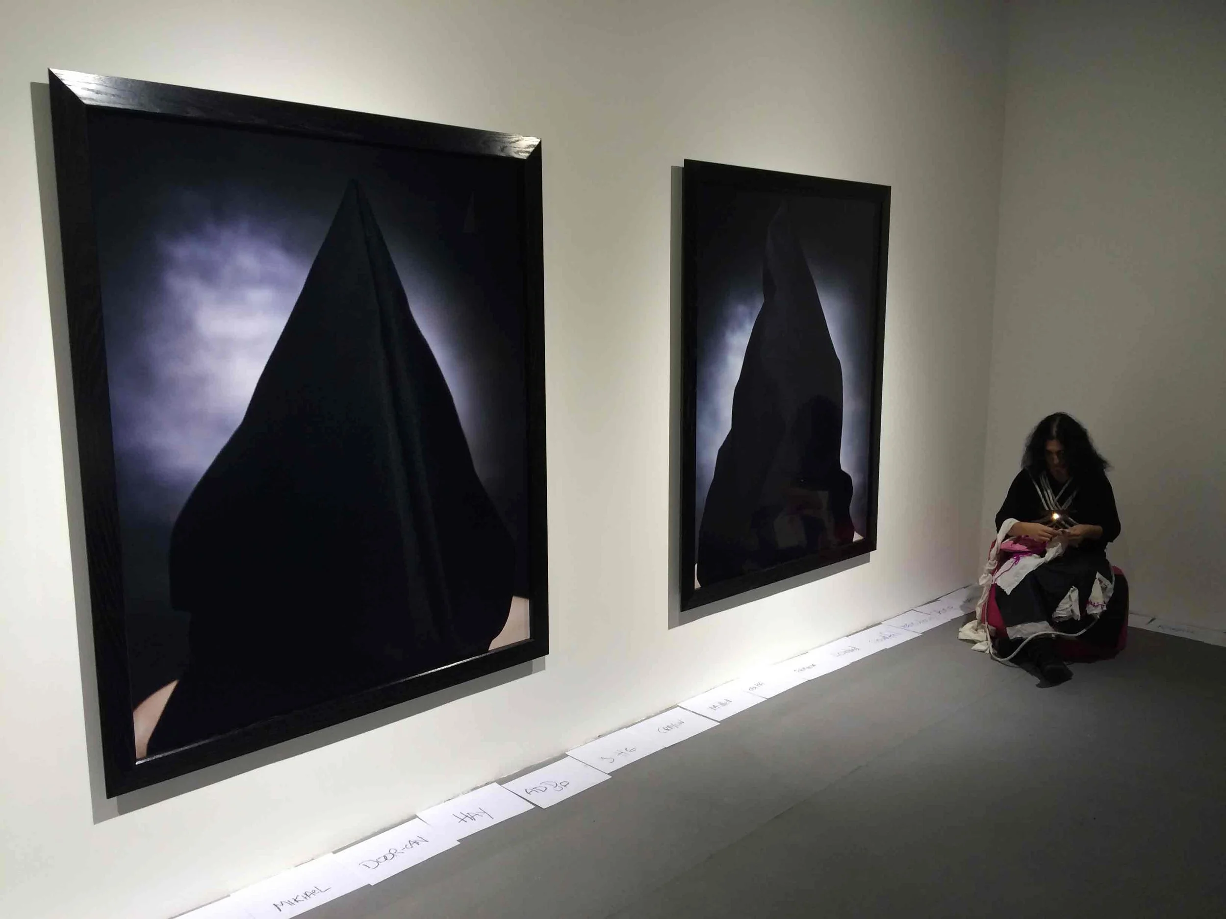 Art gallery with two large framed dark artworks on a white wall, and a person sitting on the floor with a backpack near the wall.