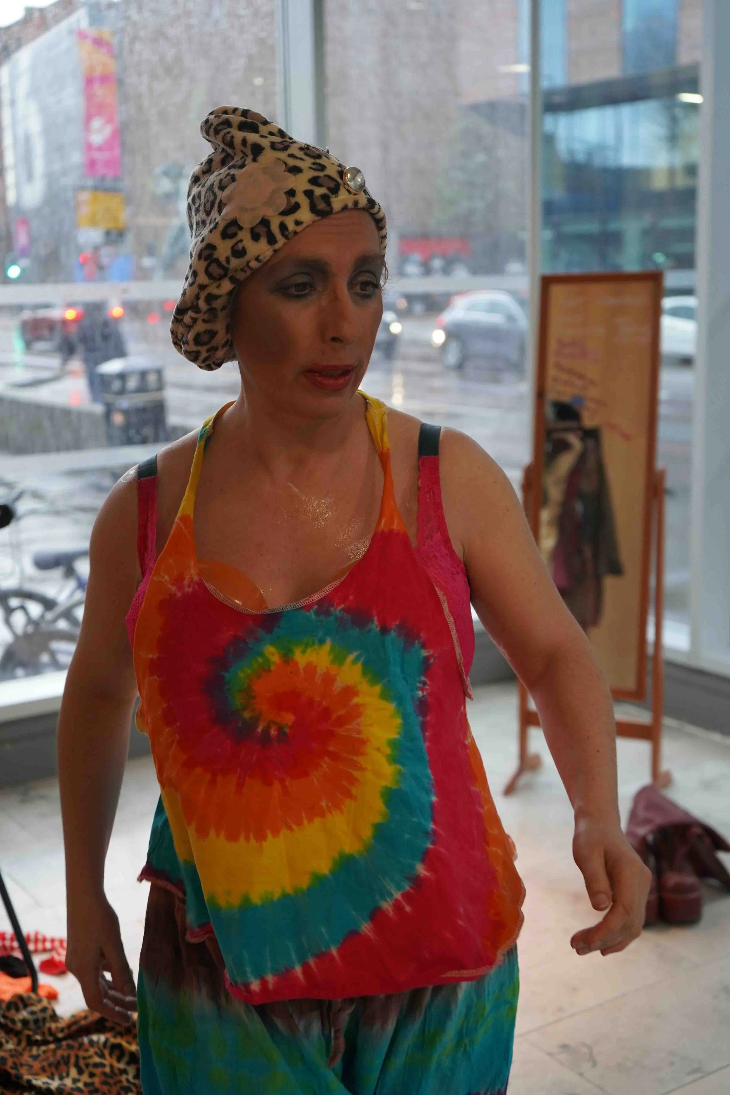 A woman wearing a colorful tie-dye top and matching pants, with a leopard print headwrap embellished with a flower and pearl detail, standing indoors near a window with a city street view in the background.