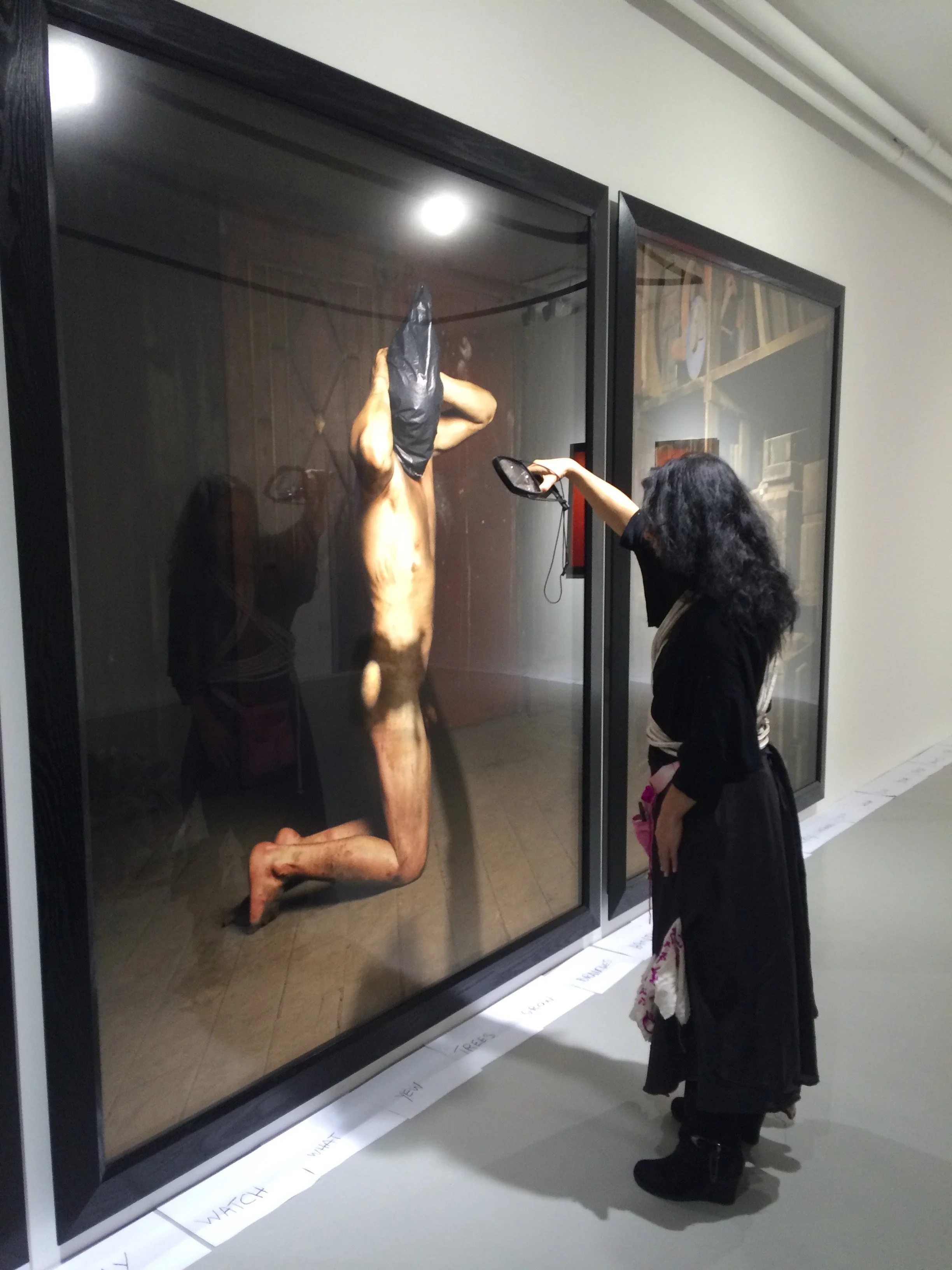 A young girl standing in front of a photograph of a naked person on their knees with a sack o n their head. The girl is wearing a dark coat and boots, and is looking at the exhibit in a gallery setting.