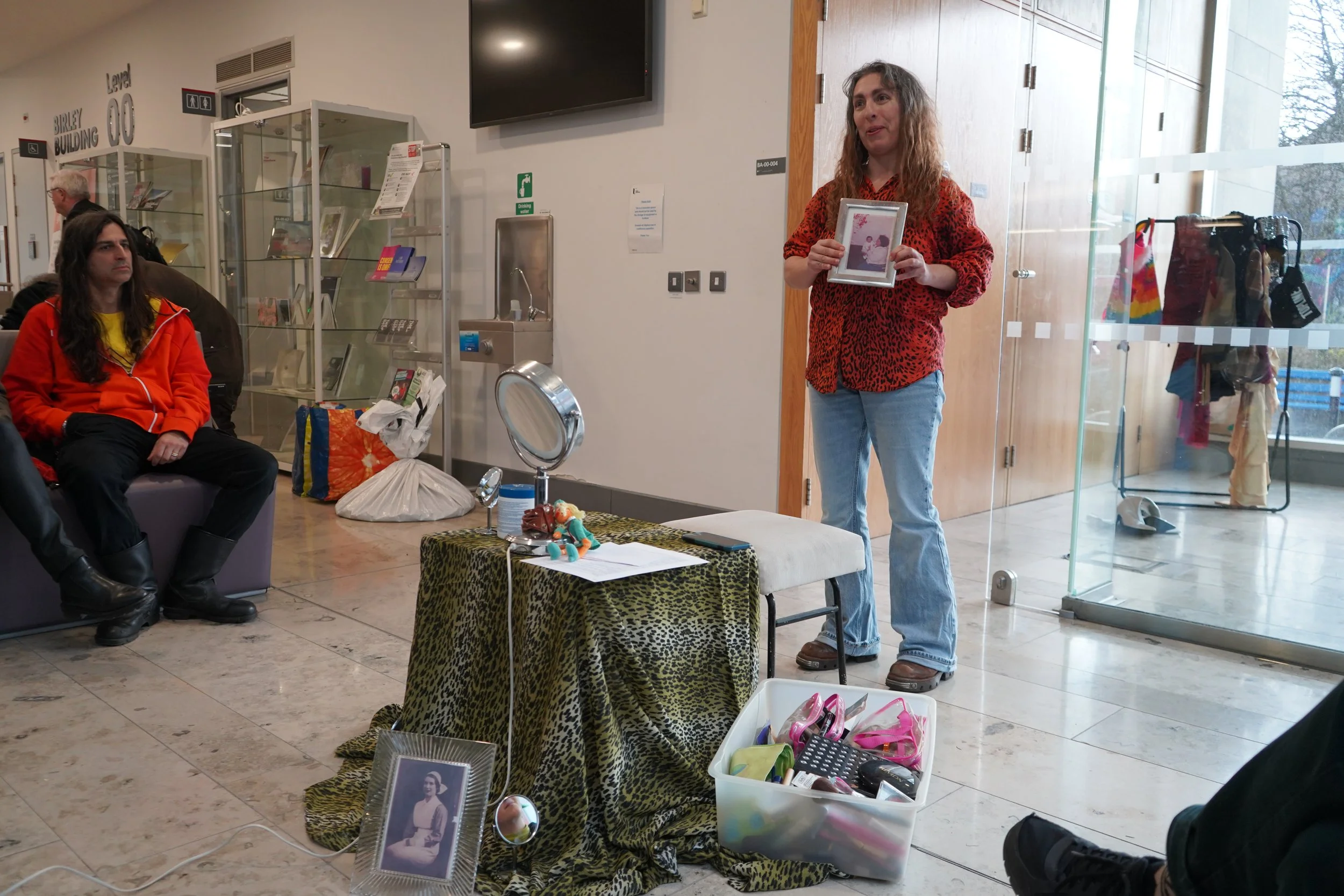Woman standing indoors holding framed photos, with clothing and accessories displayed nearby, and two seated individuals in the background in a public space such as a waiting area or community center.