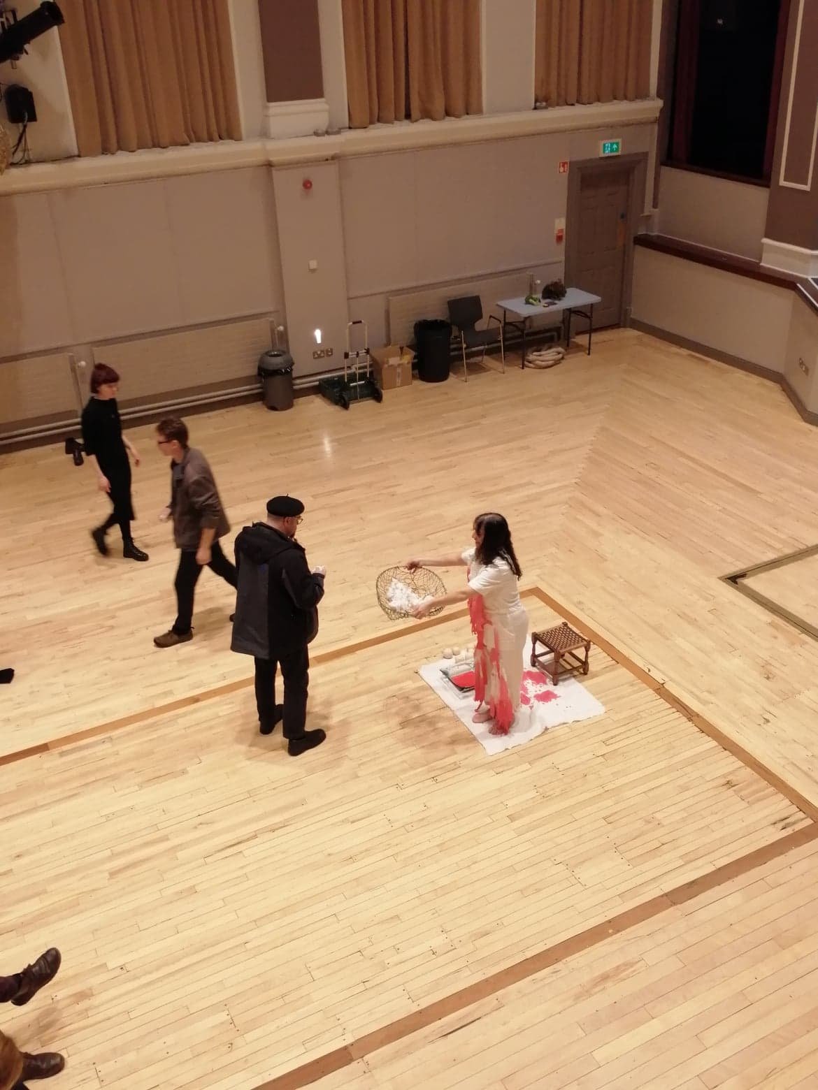 A woman in white and pink traditional attire performing a ritual on a white cloth with offerings on a wooden floor, while three men and one woman observe. The venue is a spacious hall with wooden floors, beige walls, and curtains.