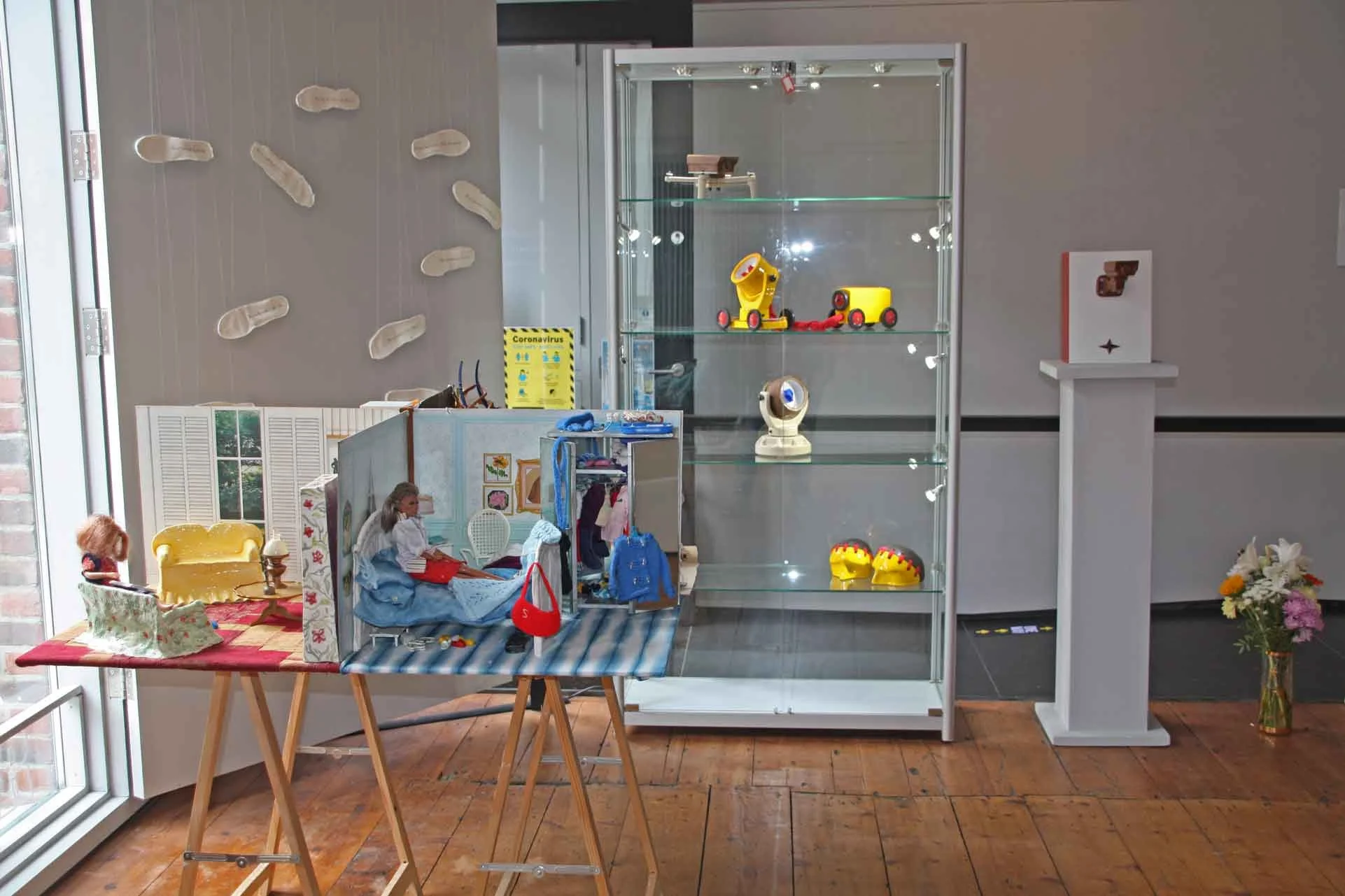 A display case with various vintage toys including a yellow toy car, a beige rotary telephone, and a small knight's helmet. To the left, a mannequin dressed in vintage clothing sits on a stool. Nearby, a table with a red tablecloth holds a floral hat and a small lamp. Behind the display, a wall features plush slippers, and a sign about coronavirus precautions. The scene is indoors with wooden flooring and large windows.