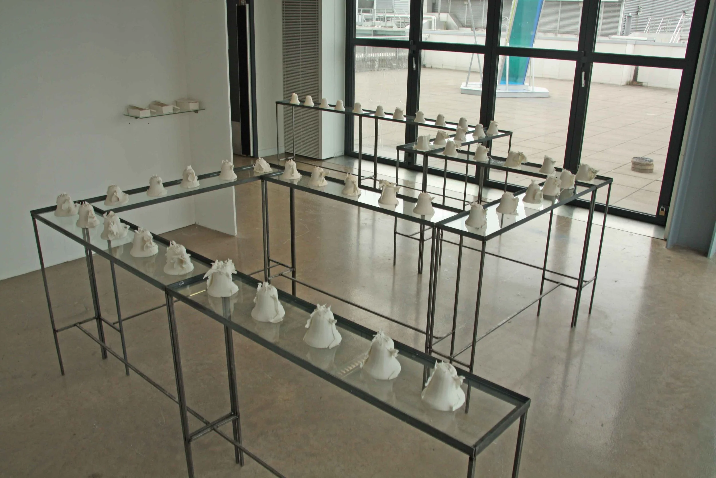 An art installation in a gallery featuring multiple white sculptures of what appears to be animal heads, arranged on black metal frames near large glass windows.