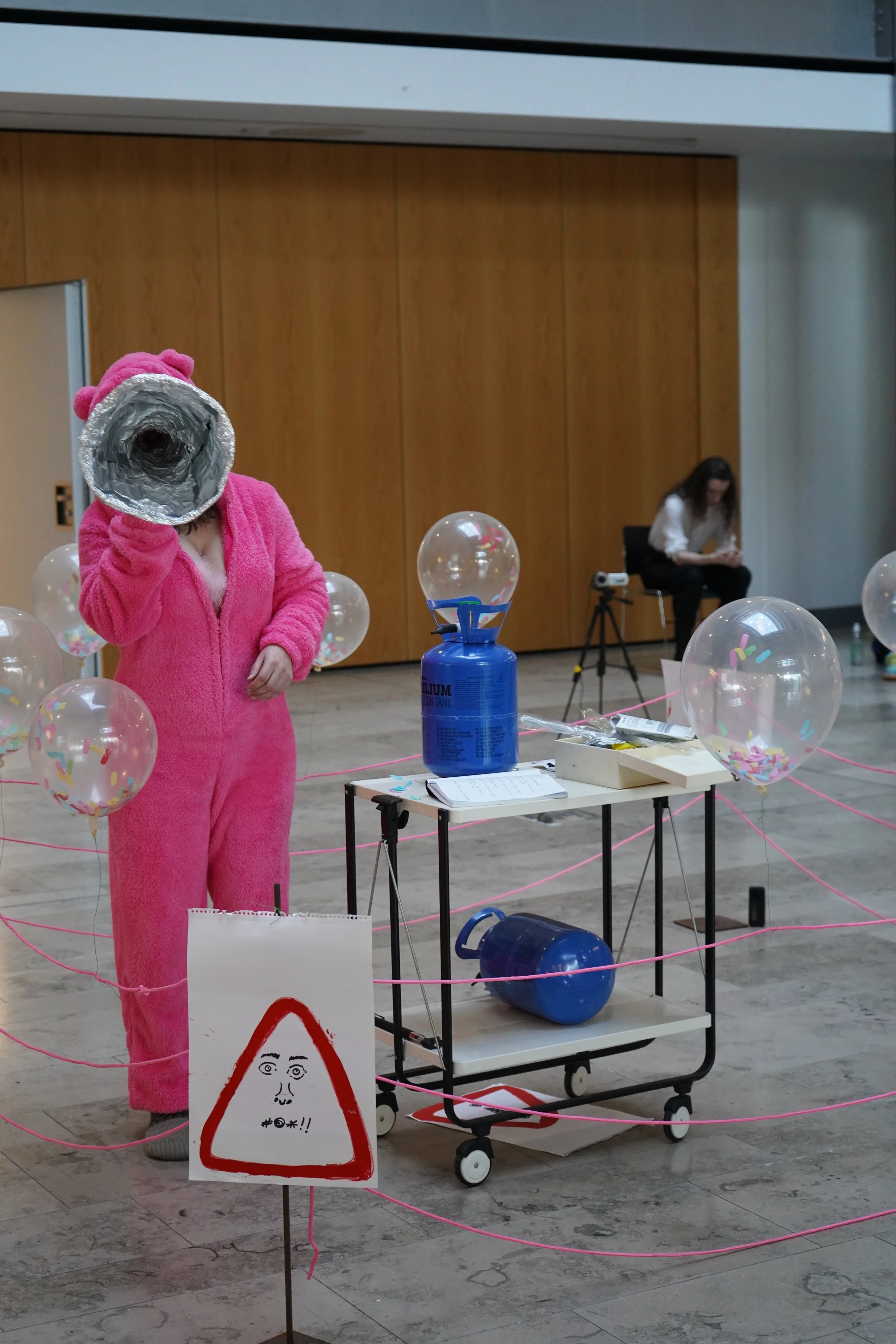 A person dressed in a pink fuzzy costume, holding a rolled-up paper tube to their face, standing in a room decorated with balloons and pink string. There is a sign with a face drawing and a warning symbol in the foreground, and a woman sitting on a bench in the background.