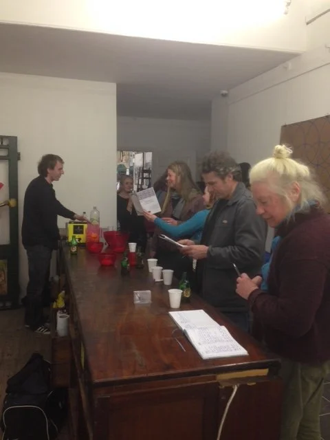 People standing at a wooden counter, reading papers, in a casual indoor setting, with some cups and bottles on the counter.