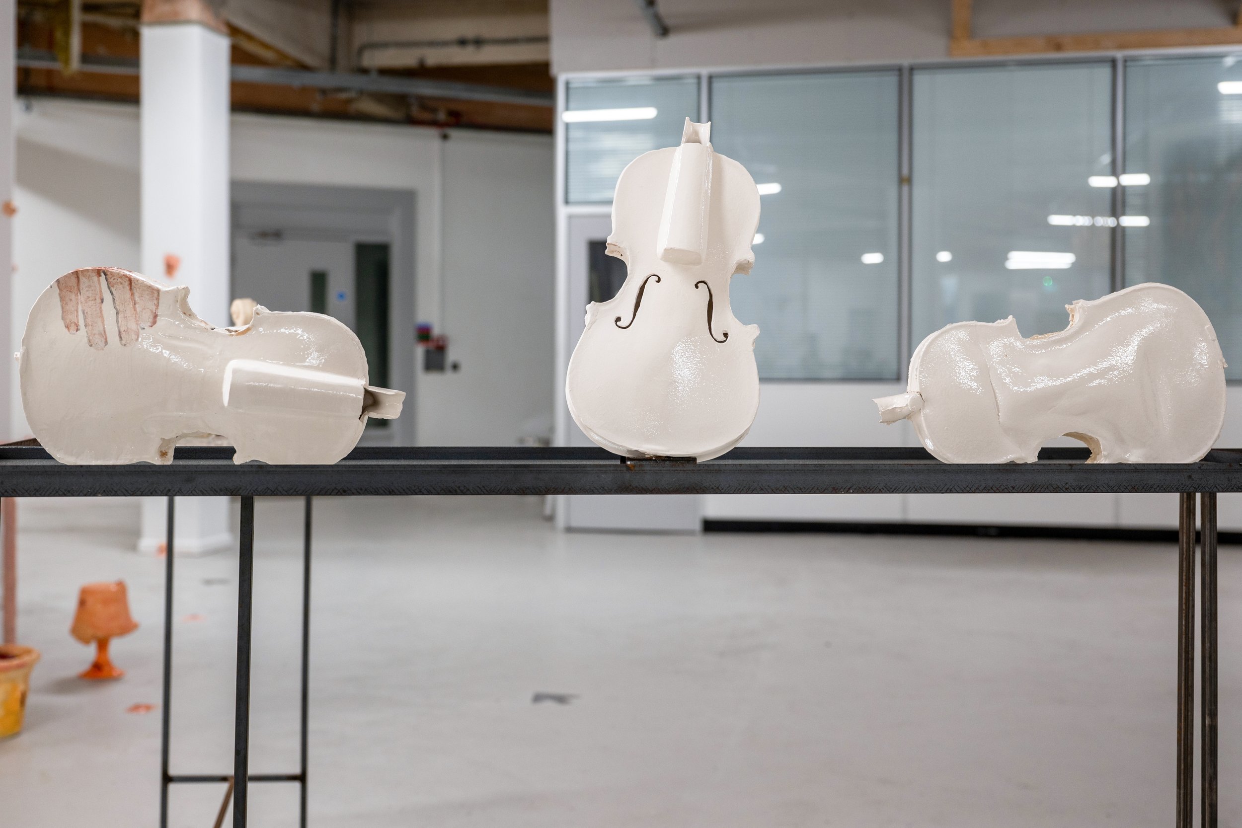 Three white, ceramic violins are displayed on a  glasstable in a modern indoor space with white walls and glass panels.