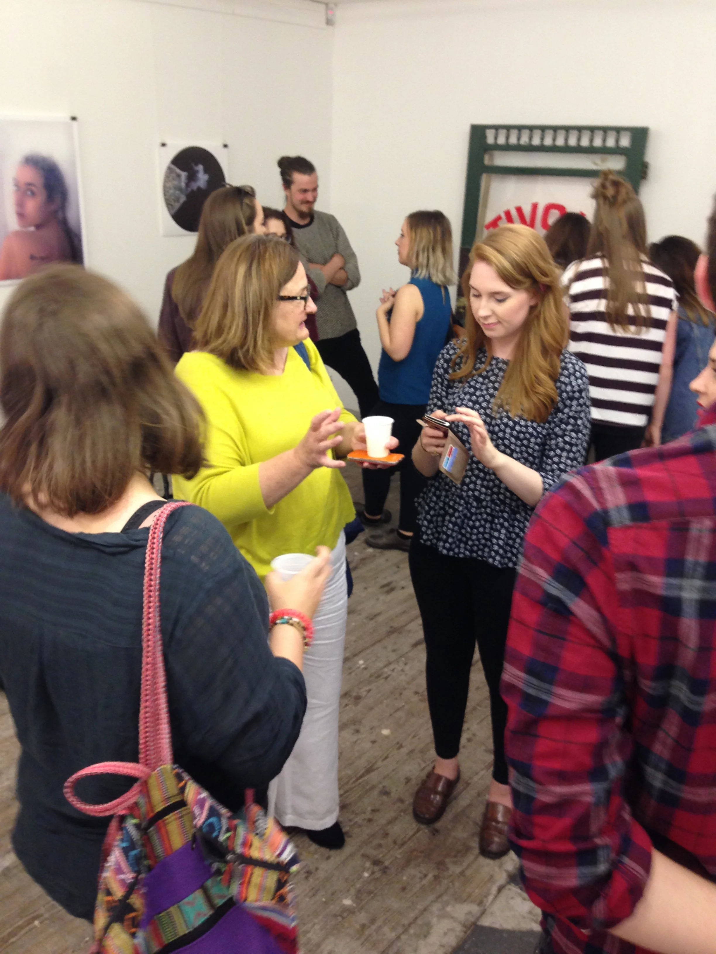 Group of people socializing at an indoor art gallery or event, with photographs on the wall and wooden flooring.