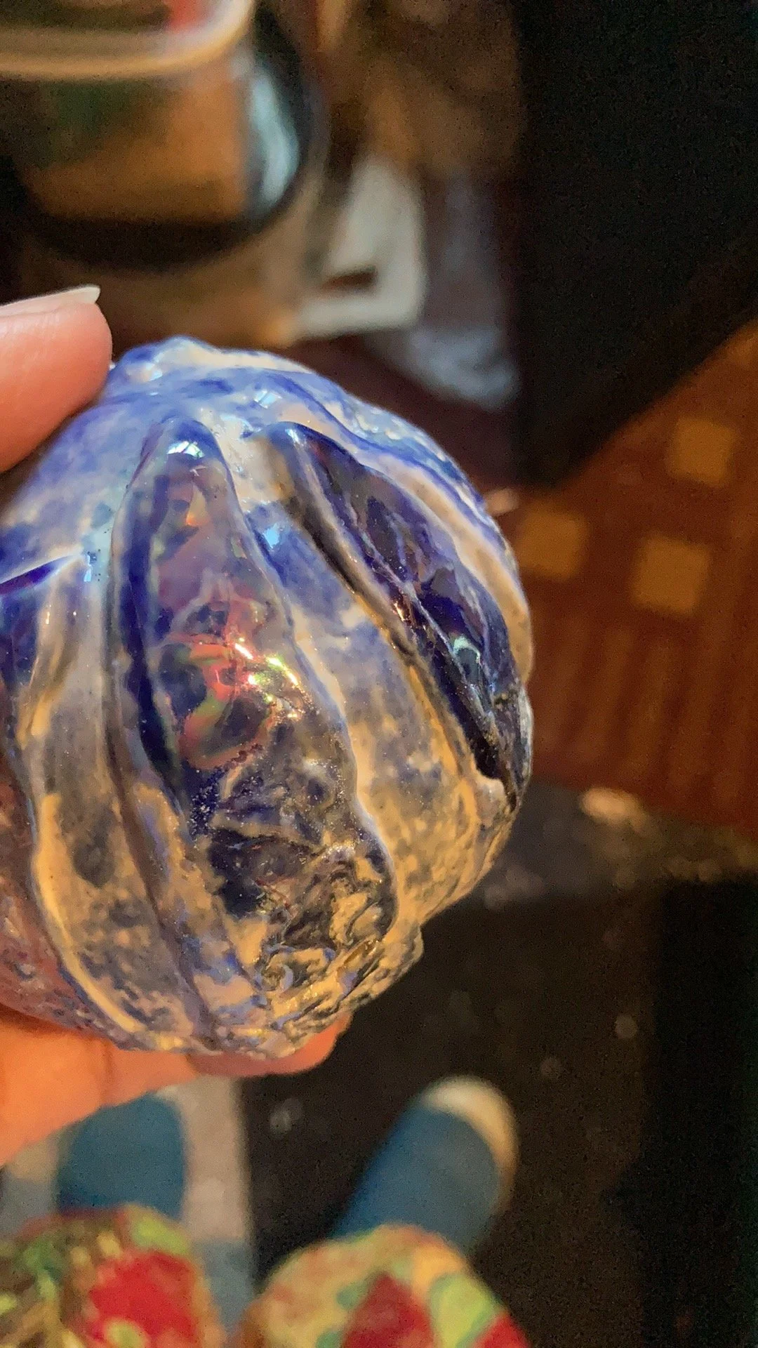 Close-up of a blue and purple glass art piece resembling a banana, being held by a person's hand, with a person's hand and colorful clothing visible in the foreground, and a table with a mug or container in the background.