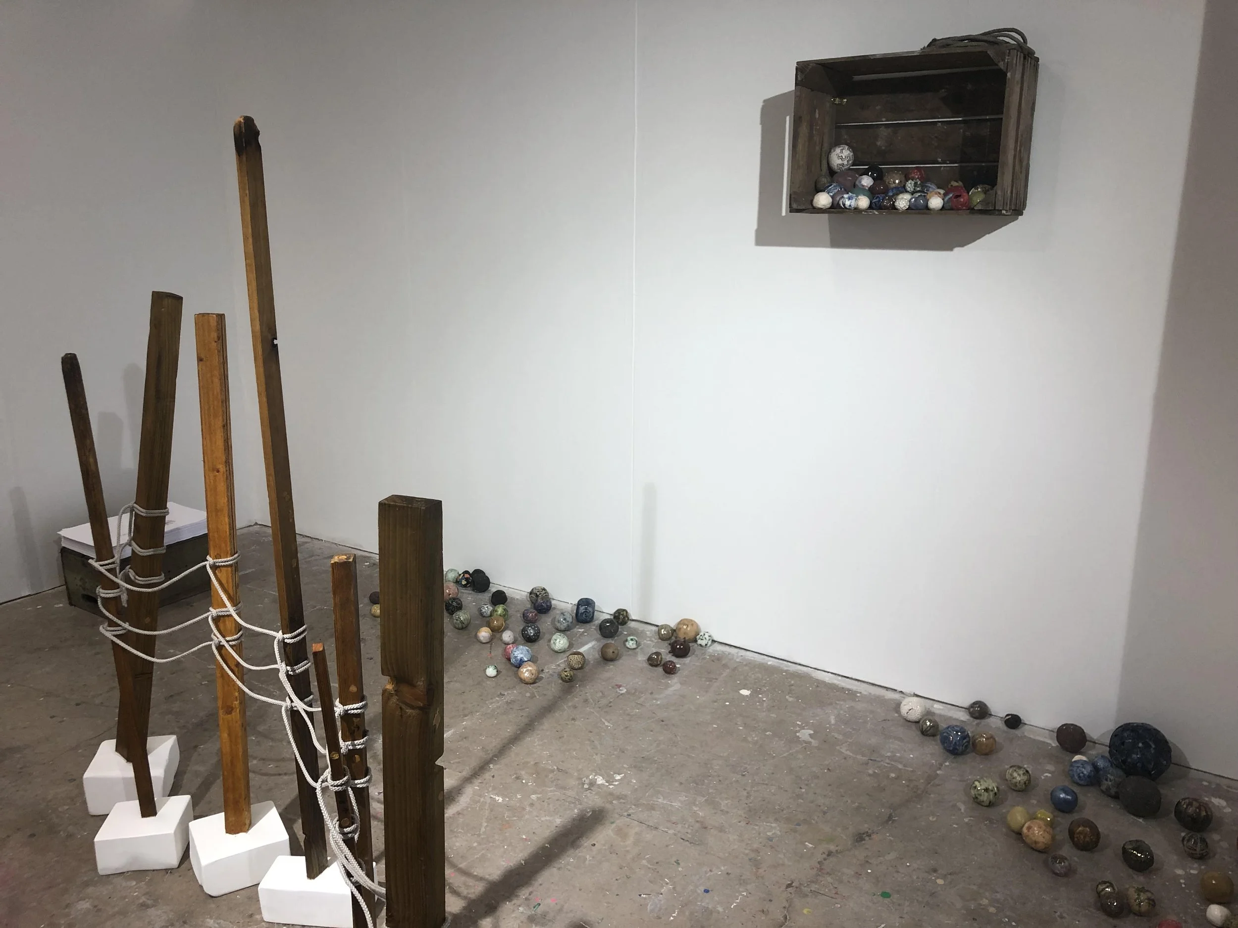 Indoor art installation with wooden structures tied together with ropes, a scattered collection of colorful marbles on the floor, and a wooden crate shelf mounted on a plain white wall.
