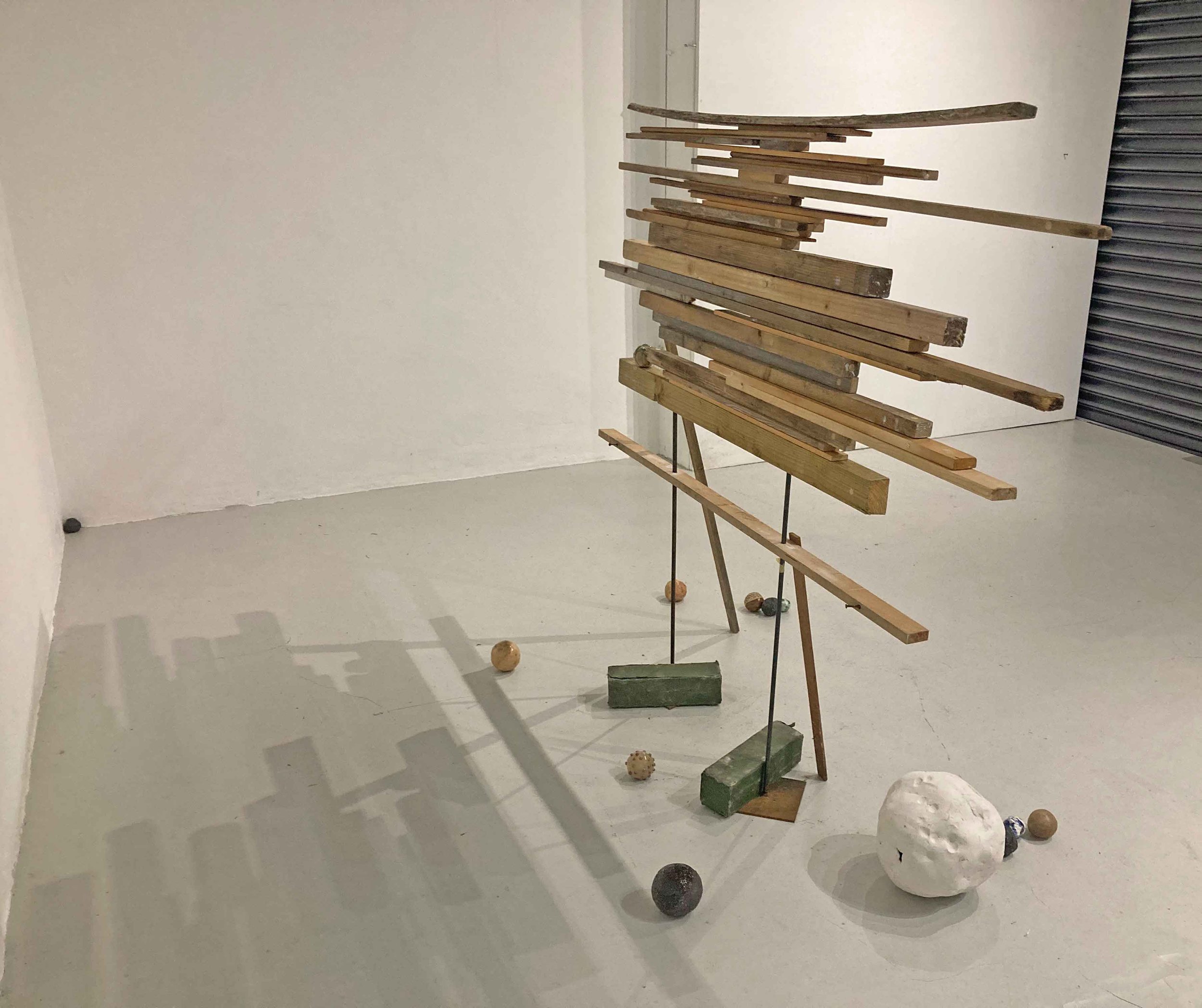 Abstract wooden sculpture with various wooden planks arranged horizontally, supported by metal rods, with gravel balls on the floor.