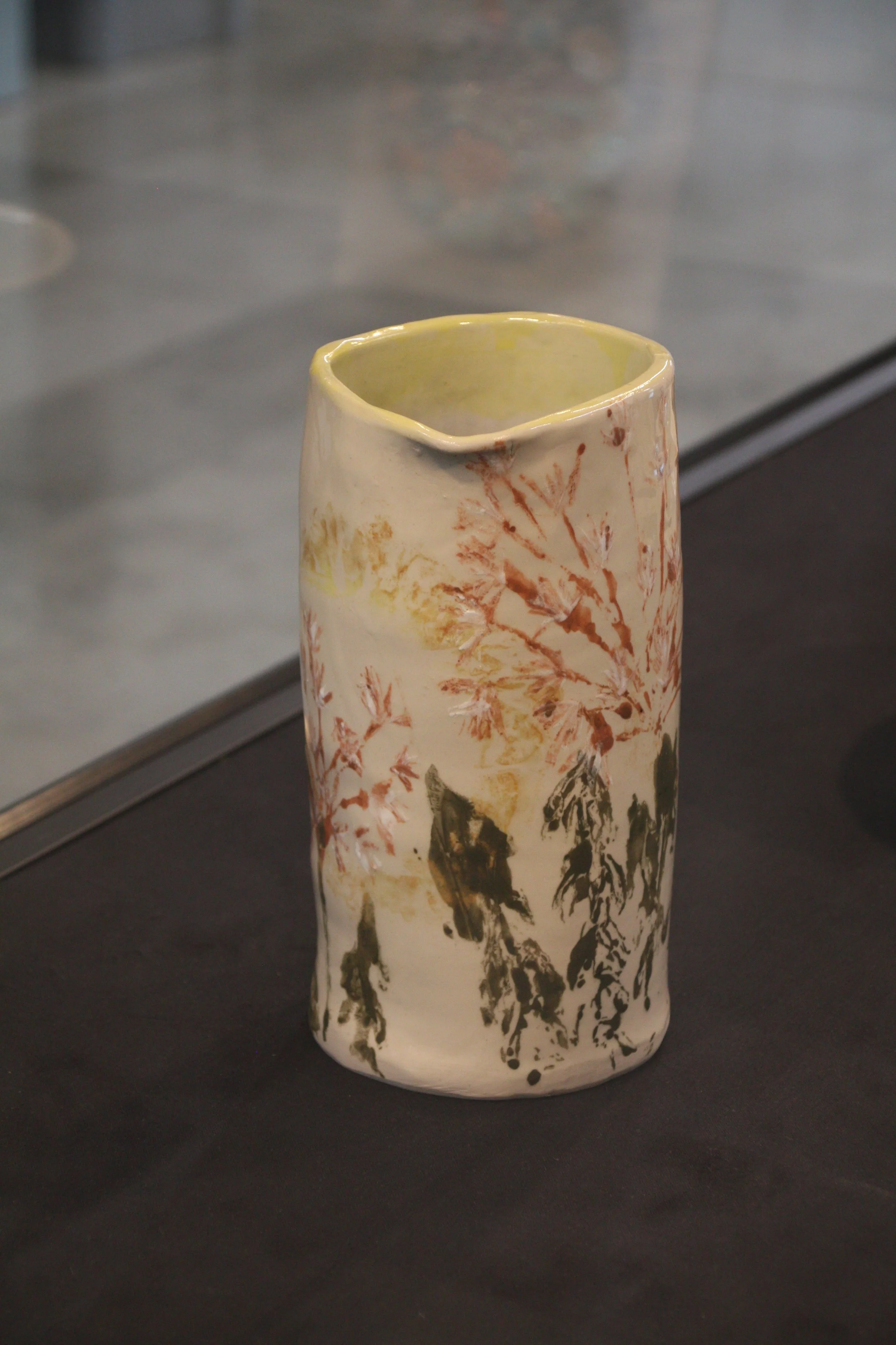 A tall ceramic vase with a narrow opening at the top, decorated with abstract painted designs in earthy tones of brown, black, and red, and a background of white glaze, displayed on a dark surface.