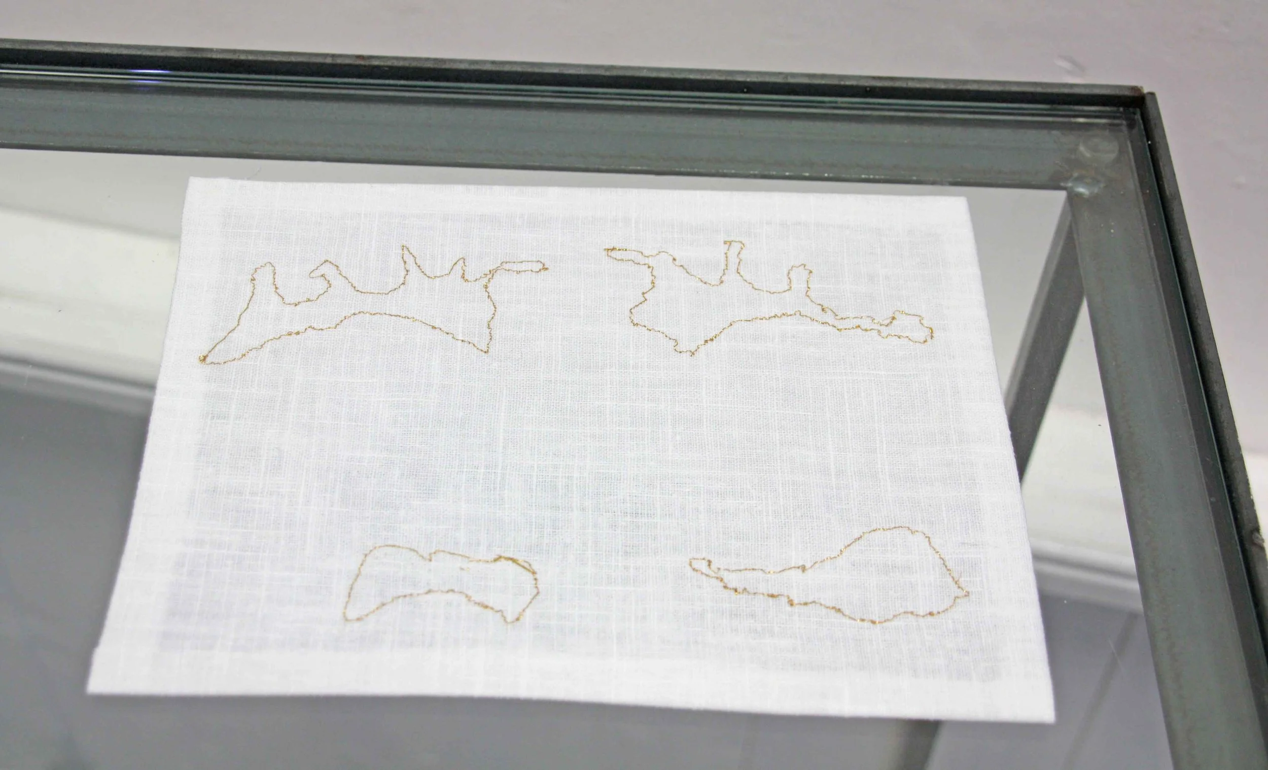A piece of white fabric with gold embroidery outlines of various shapes inside a glass display case.