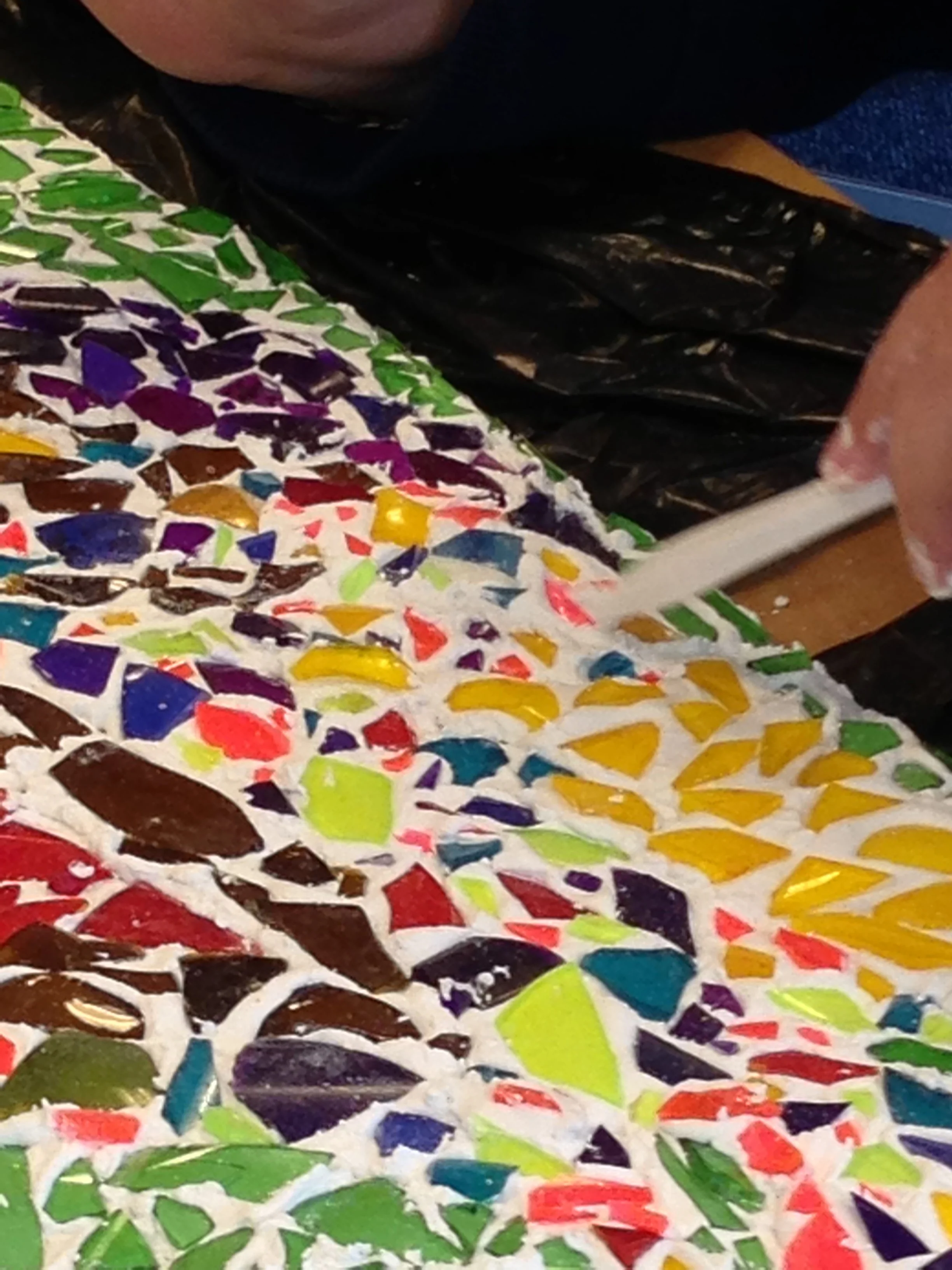 Close-up of a colorful mosaic made of small, irregularly shaped pieces of colored glass or ceramic, including green, red, yellow, purple, blue, and brown.