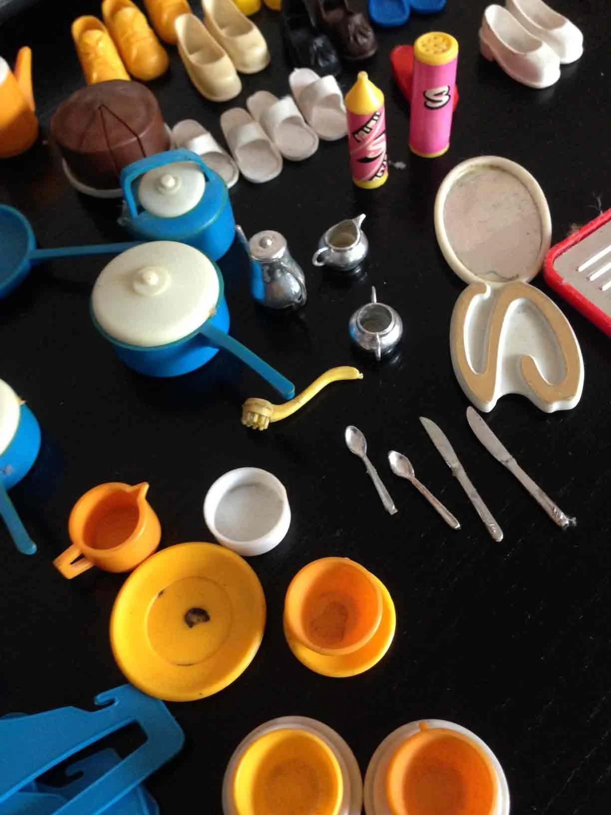 A collection of toy kitchen items including pots, pans, utensils, cups, and butter knives, arranged on a black surface.