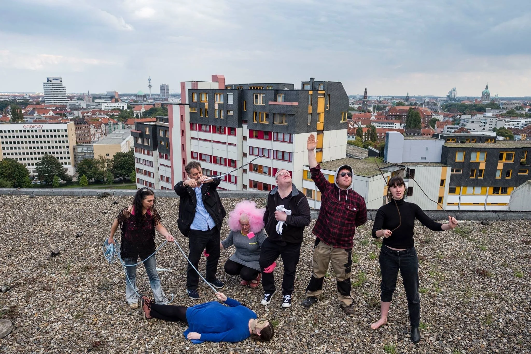 Group of people participating in an outdoor skit or performance on a rooftop with city skyline in the background, some holding ropes and one lying on the ground as if playing a dead body.