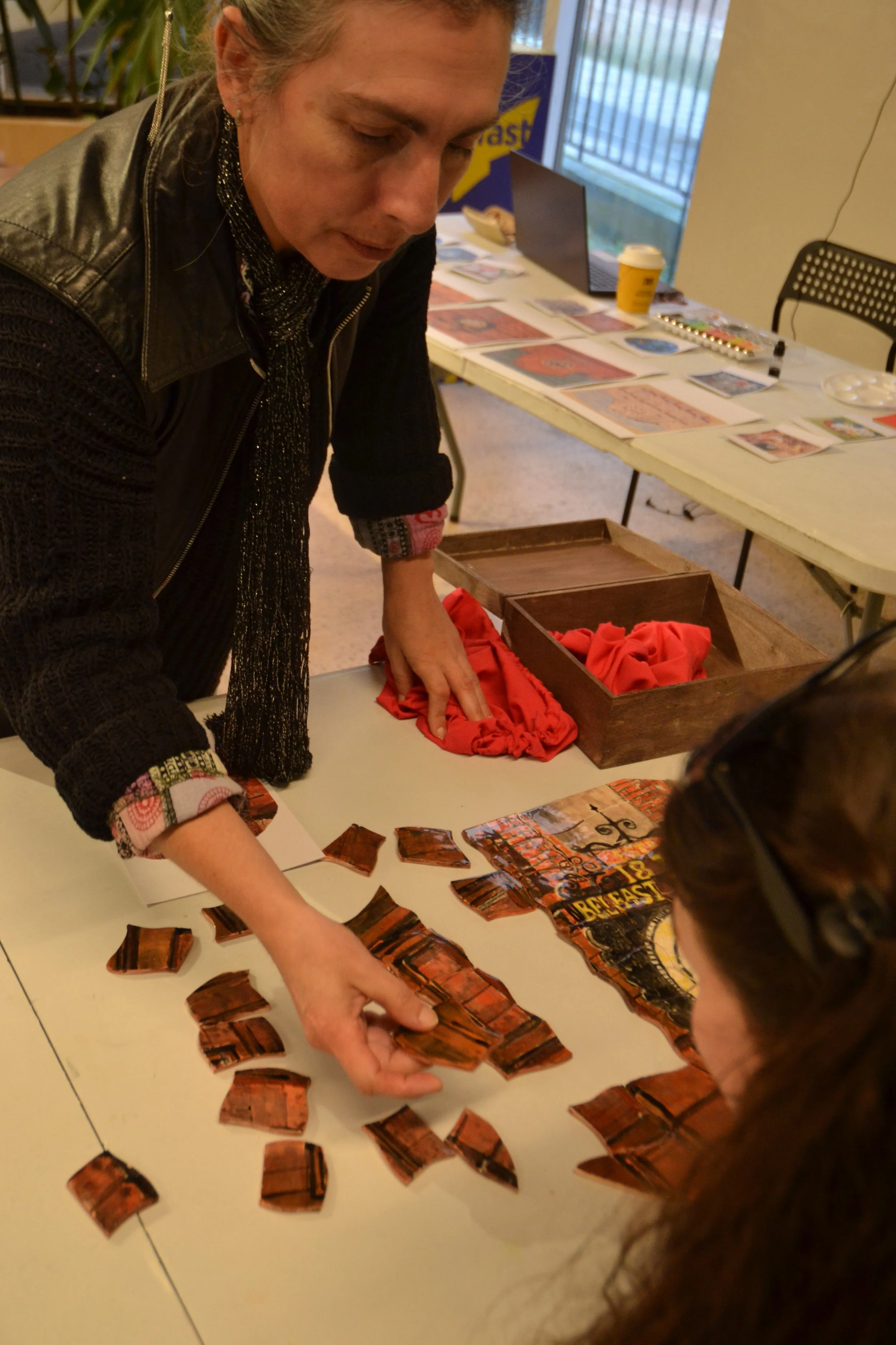 People working on a puzzle spread out on a table, with some puzzle pieces already assembled. Background includes a laptop, a cup, and printed materials on the table.