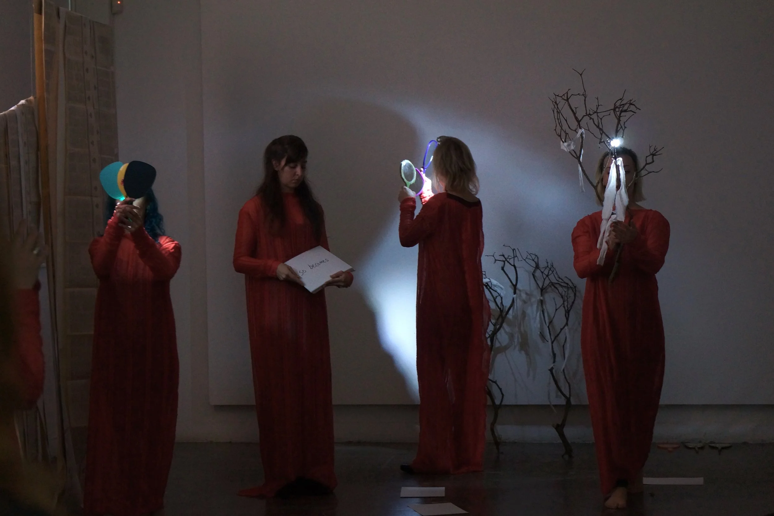Four people dressed in red robes standing in an indoor gallery. One person on the far left is holding colorful ping pong paddles and a ball. The second person from the left is holding a notebook with writing that says 'so becomes'. The third person i