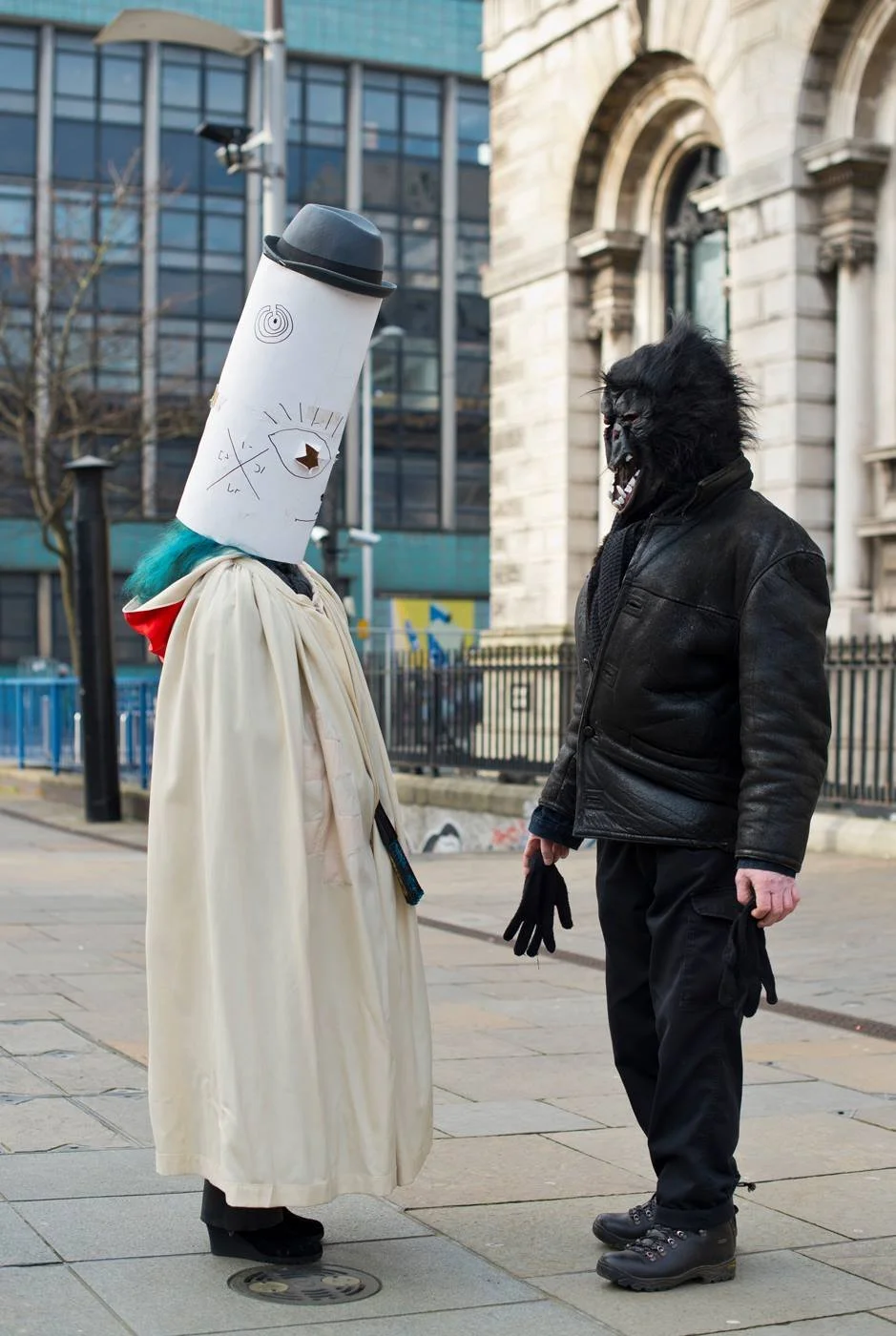 Two people in Halloween costumes face each other on a city sidewalk, one dressed as a tall paper cup with a face drawn on it and the other as a gorilla with a fierce mask, both wearing black gloves.