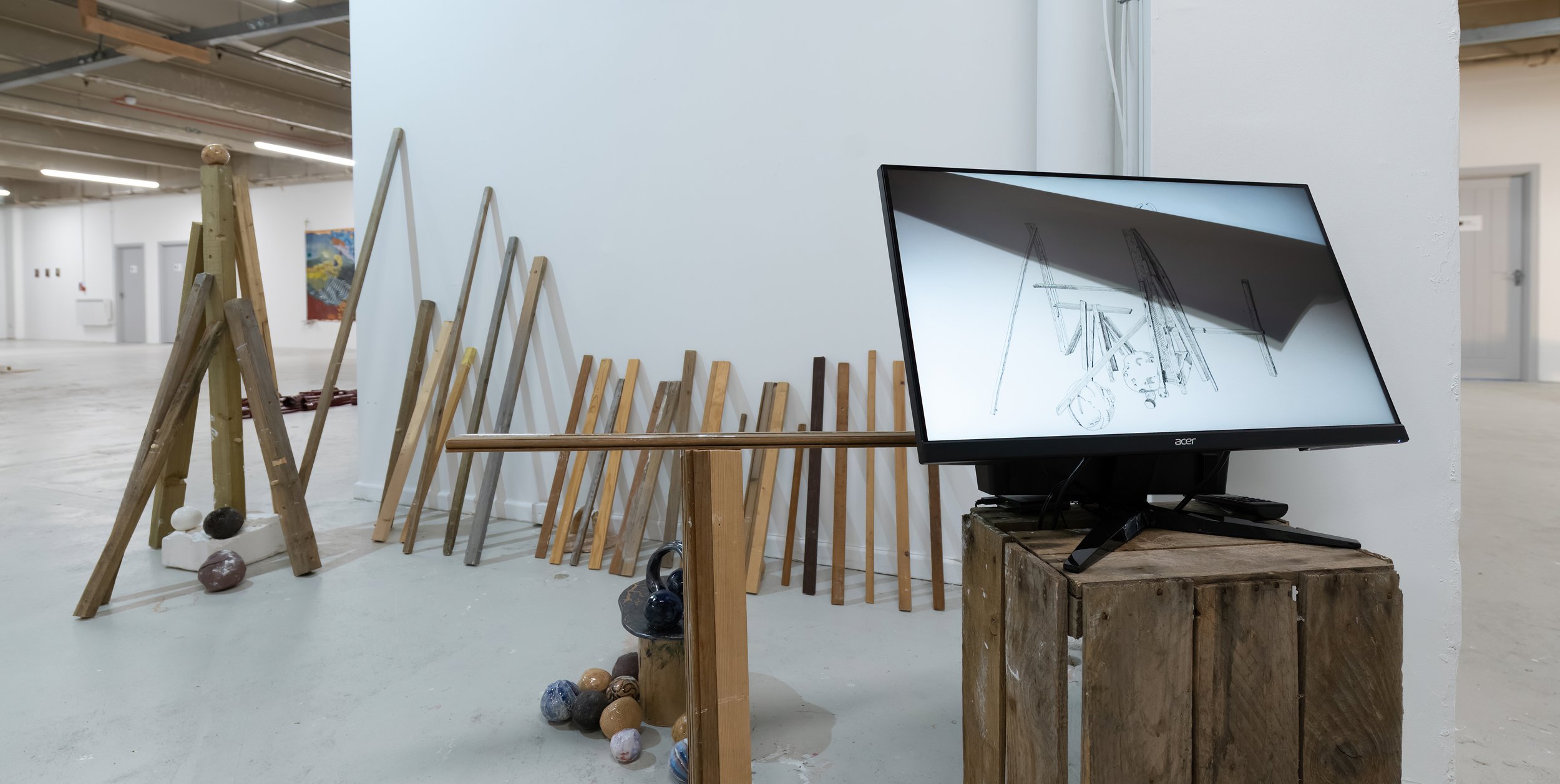 An art installation in a gallery featuring wooden sticks leaning against a white wall, a monitor displaying a sketch of construction equipment, and painted rocks on the floor.