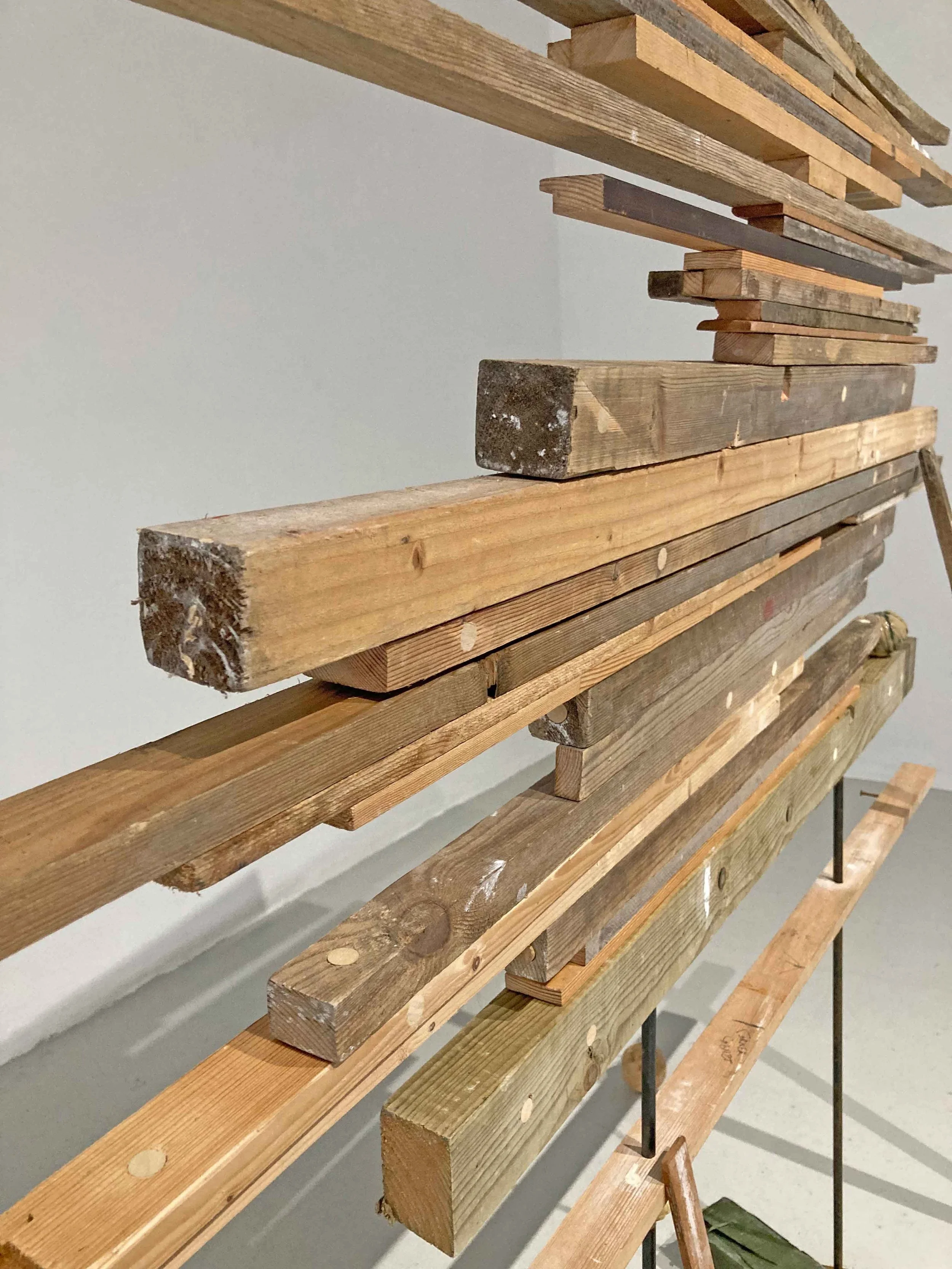 Multiple wooden planks arranged horizontally on a metal rack, with some planks showing signs of weathering and nail holes, set against a plain gray wall.