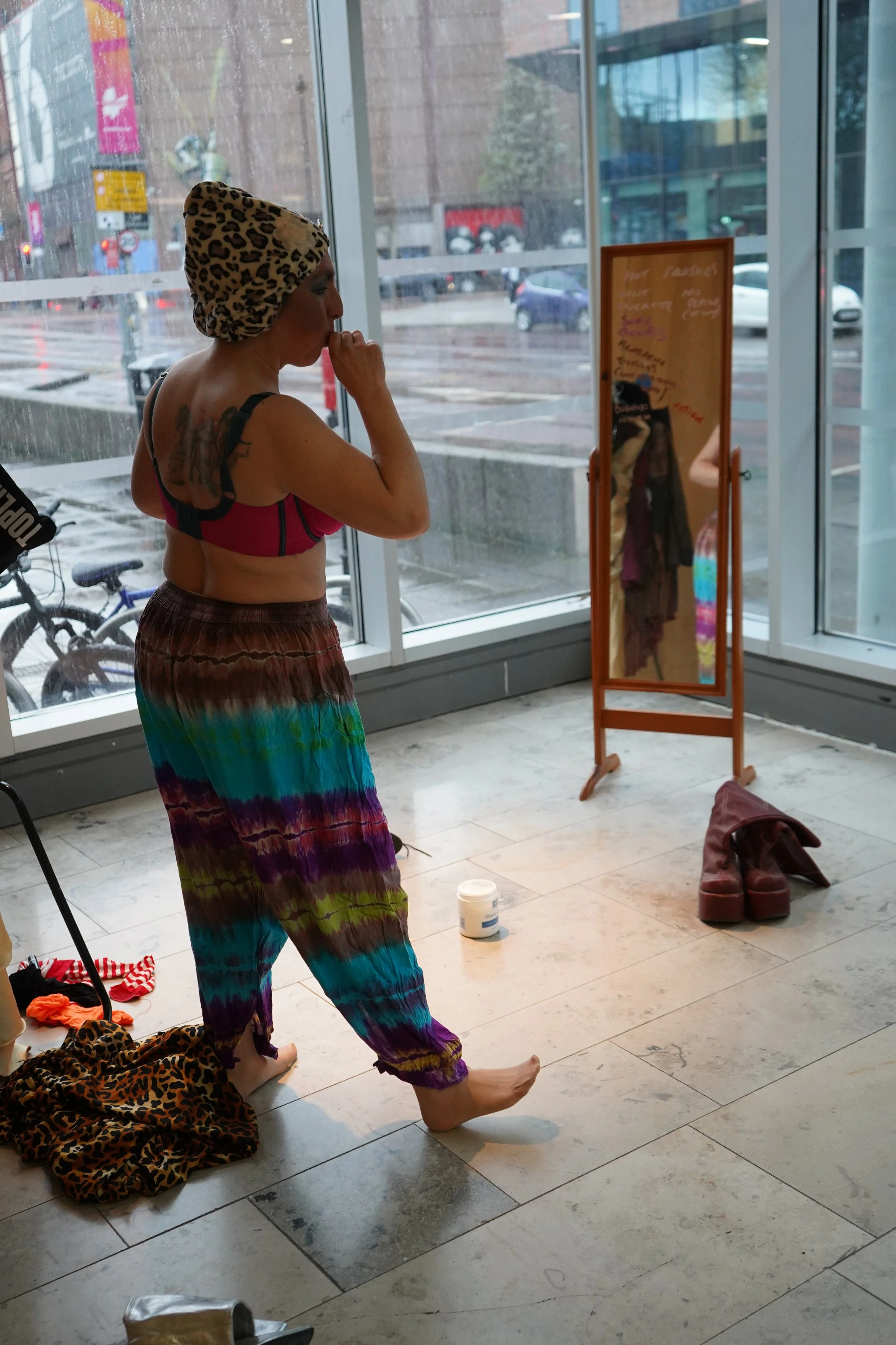 A woman with short hair, wearing a leopard print headscarf, a red bra, and rainbow tie-dye pants, standing barefoot inside a store. She appears to be preparing for a performance or dance. There are shoes, a leopard print fabric, and a mirror in the s