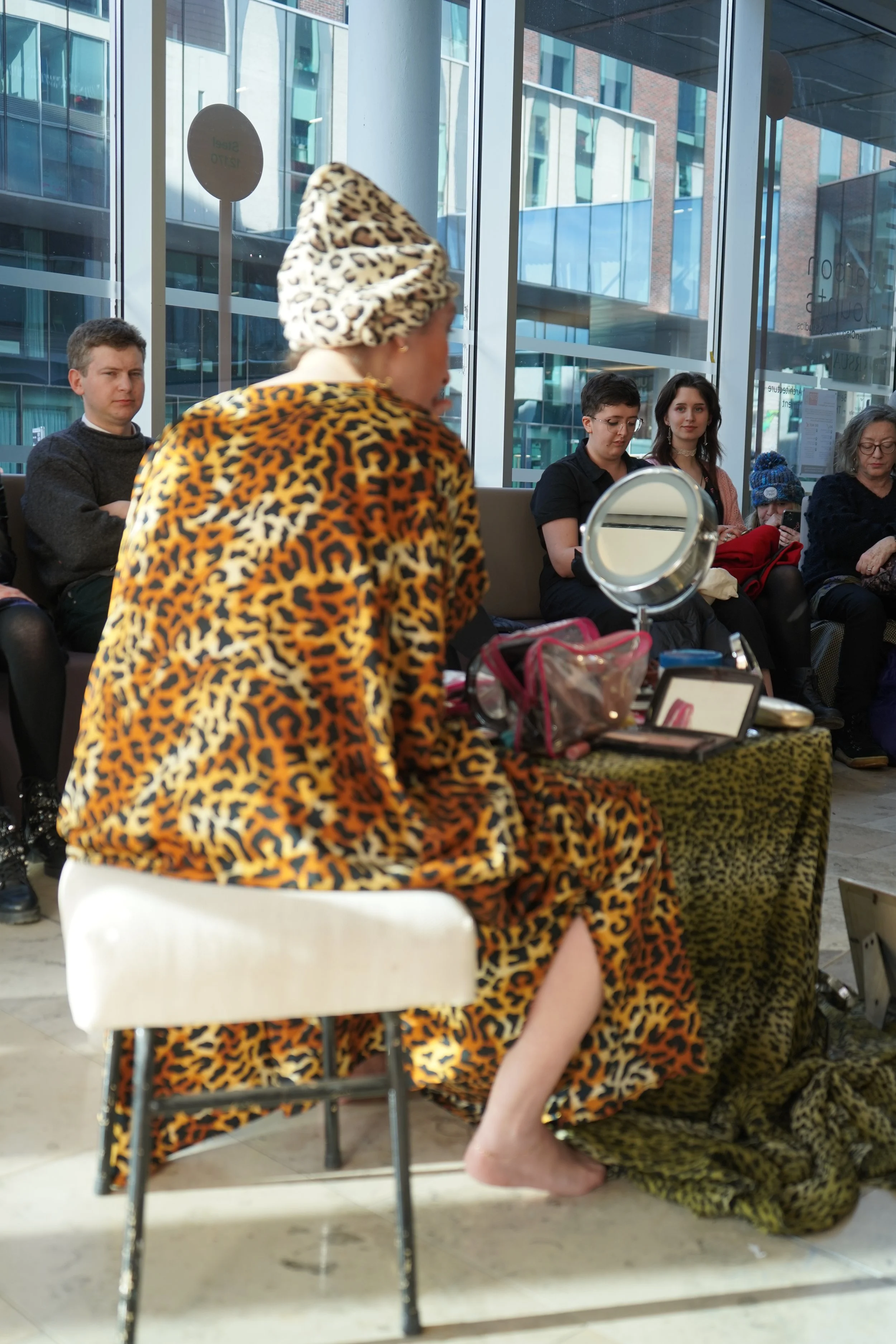 A woman wearing a leopard print coat and a leopard print hat is sitting at a makeup station inside a modern building with large windows. Several people are seated in the background, some using their phones, with buildings visible outside the windows.