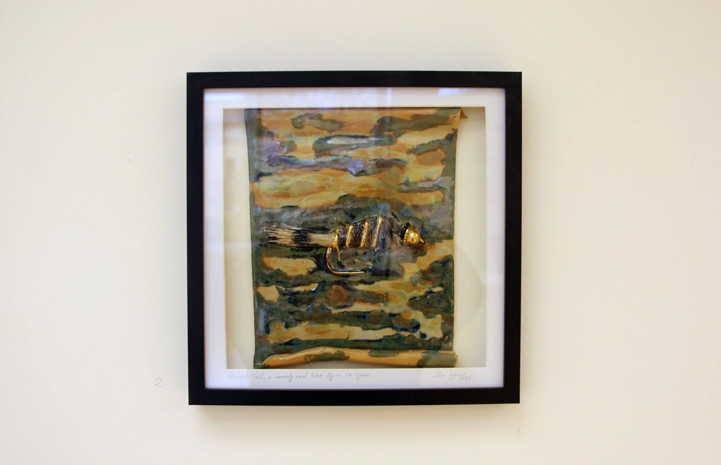 A framed ceramic fishing lure tile gold and black coloring resting on a surface at the bottom of the artwork.