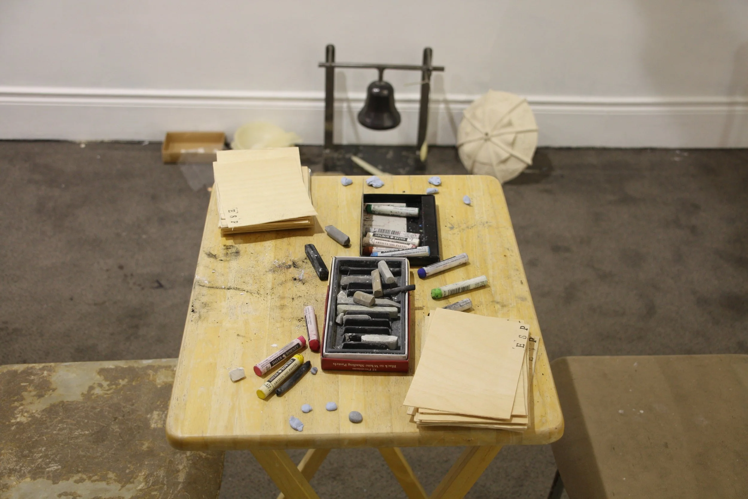 A yellow wooden table with various art supplies including pastels, chalks, and paper sheets, set against a plain wall and a gray carpeted floor.