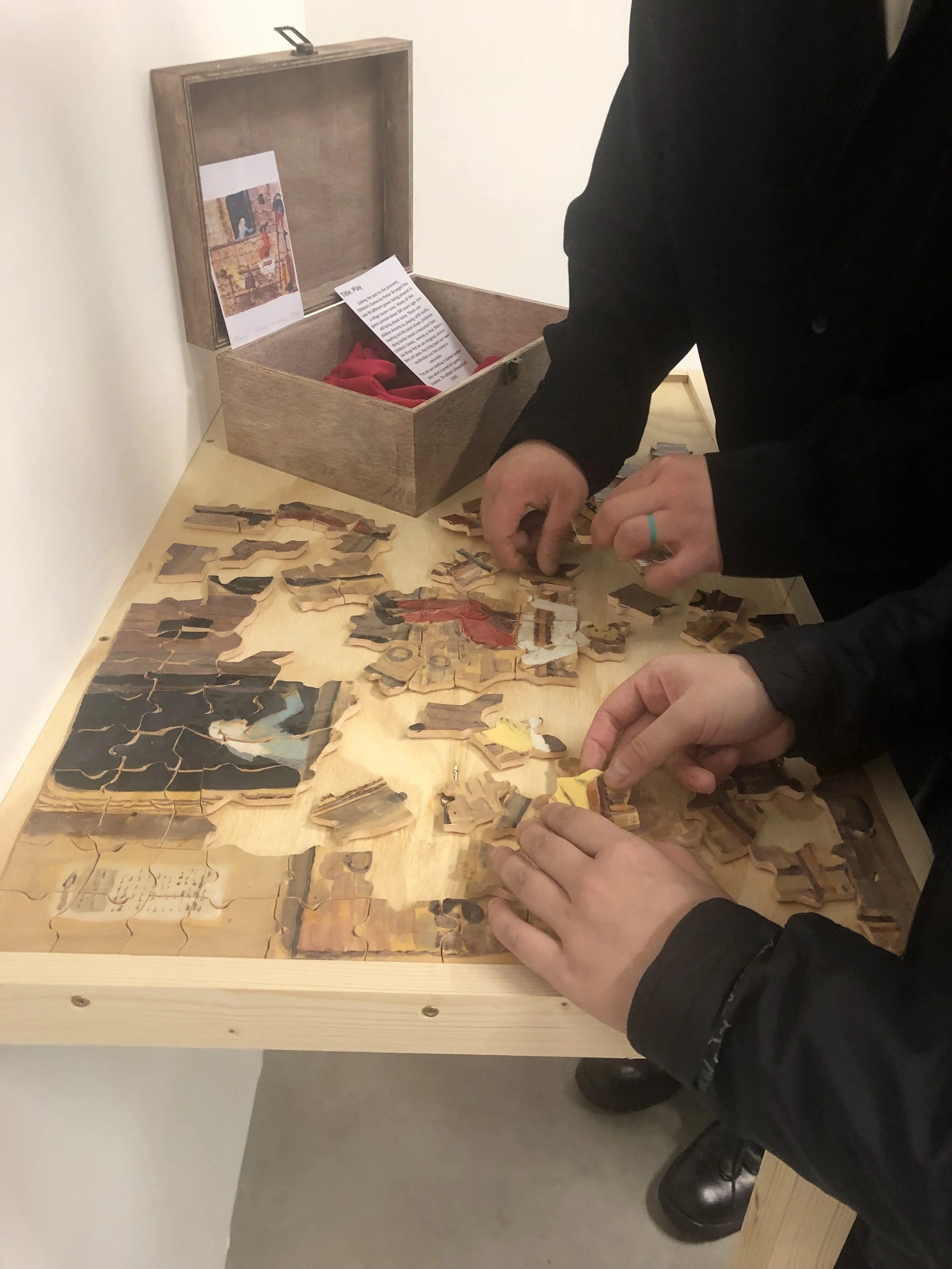 People assembling a puzzle on a wooden table, some puzzle pieces are visible, with a box and printed guide in the background.