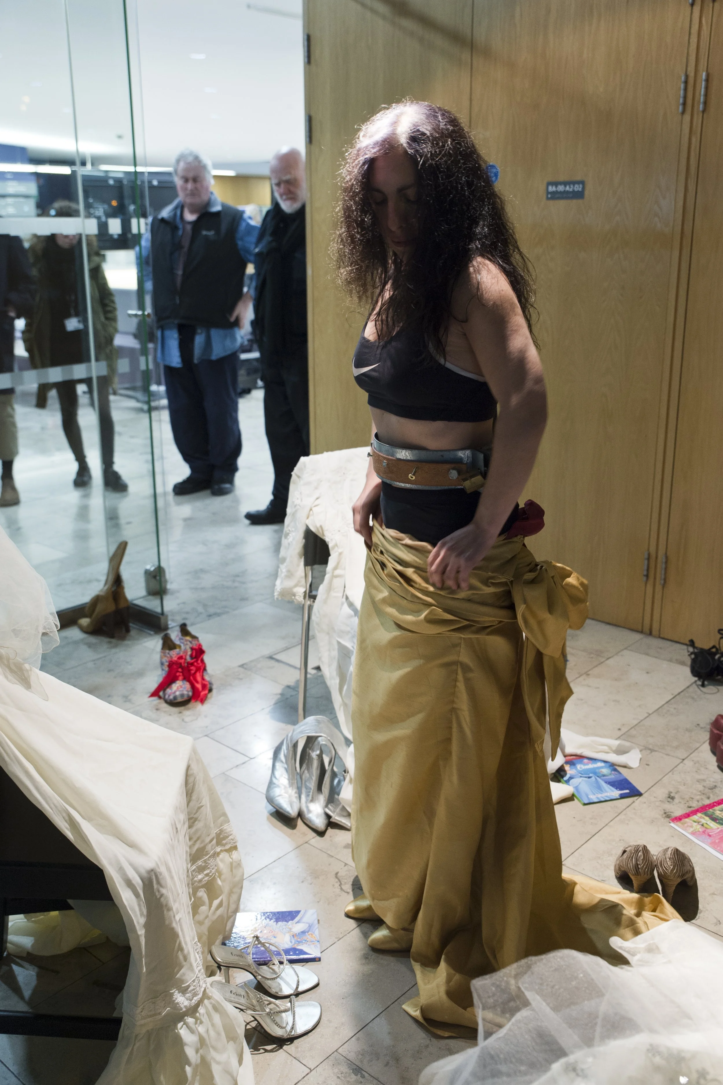 A woman with long, curly hair is looking down, dressed in a black sports bra and a long, gold-colored skirt. She appears to be trying on clothes in a store, with various shoes and clothing items scattered nearby. In the background, a glass wall and s