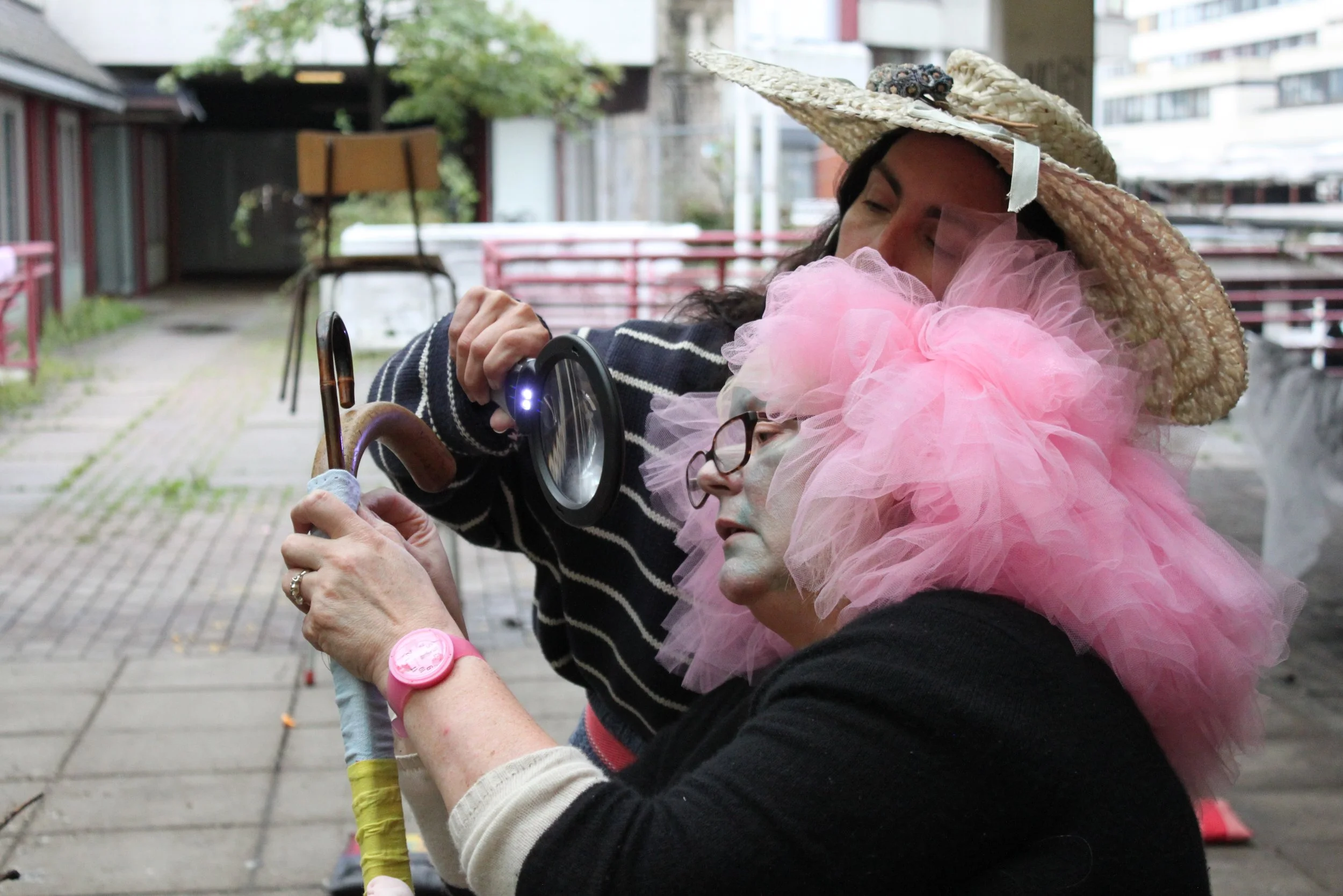 Two women dressed in costume, one with a large pink tulle tutu and the other with a straw hat adorned with a small animal figure, sharing a moment together outside on a paved area.