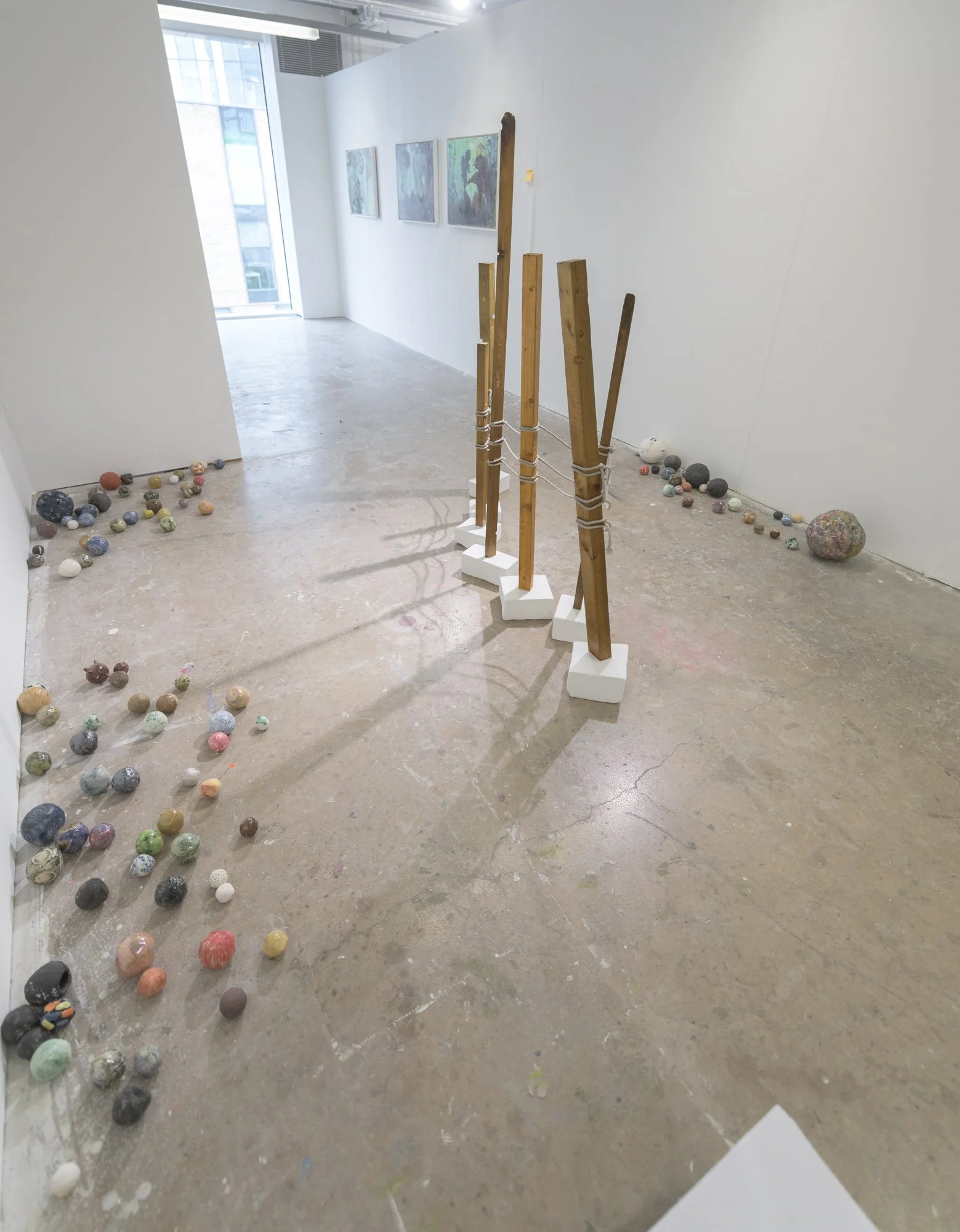 Art installation in a gallery with colorful painted stones scattered on the floor, wooden poles with string attachments, and paintings on a white wall.
