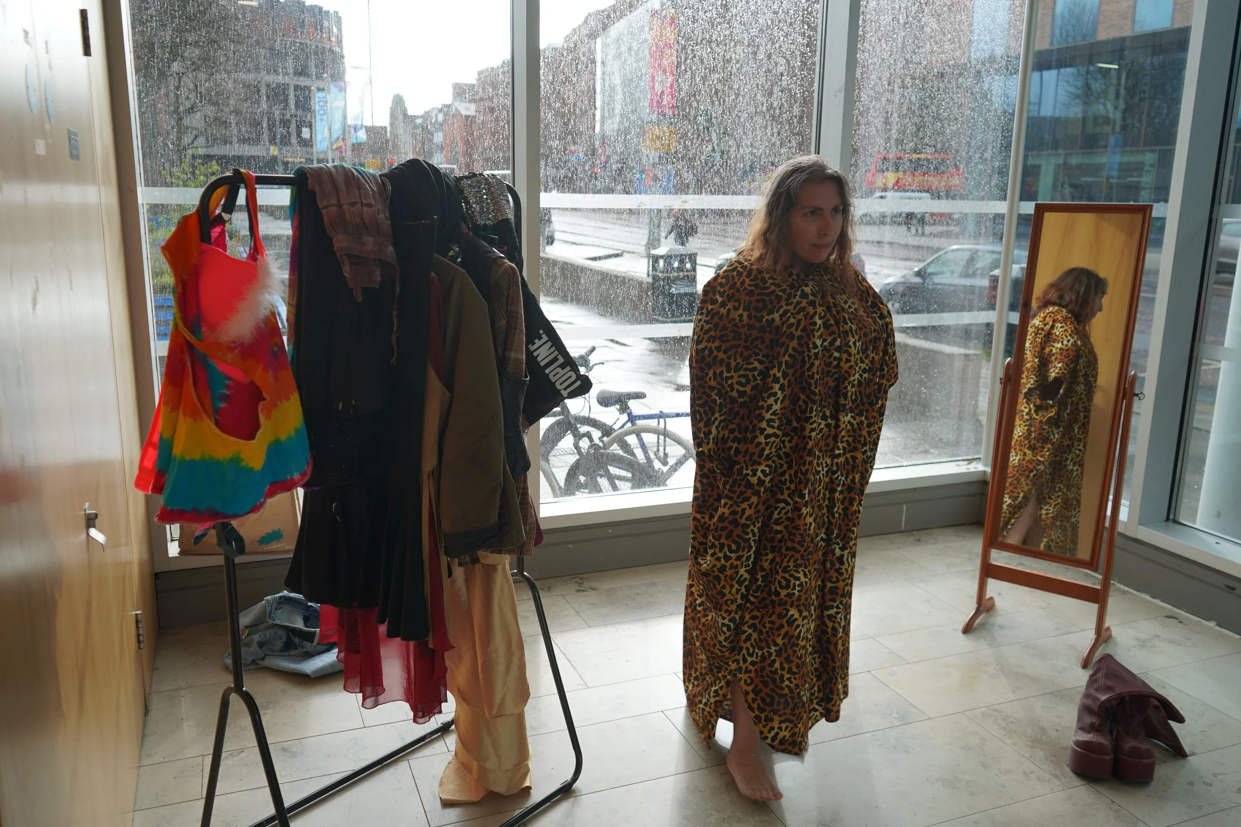 A woman with curly hair wearing a leopard print dress standing barefoot in front of a large mirror in a room with a glass window and door. Outside, it is raining with blurry reflections of city buildings and vehicles. Inside, there is a clothing rack