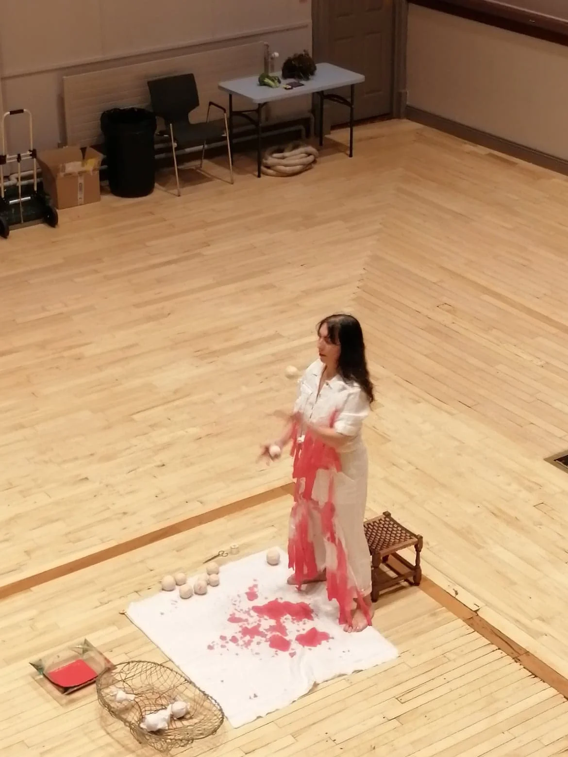 A woman standing on a white cloth covered with spilled red liquid and surrounded by ping pong balls in a large, empty room with wooden floors. She appears to be involved in a performance or art piece.